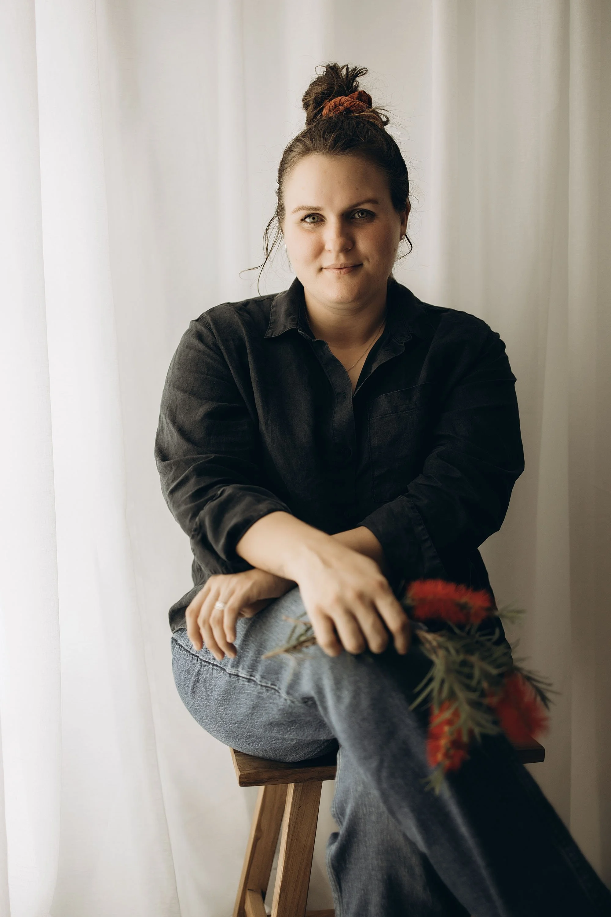A woman in a dark linen shirt sitting on a stool in a home photography studio in Darwin, holding a bunch or bright red bottle brush flowers