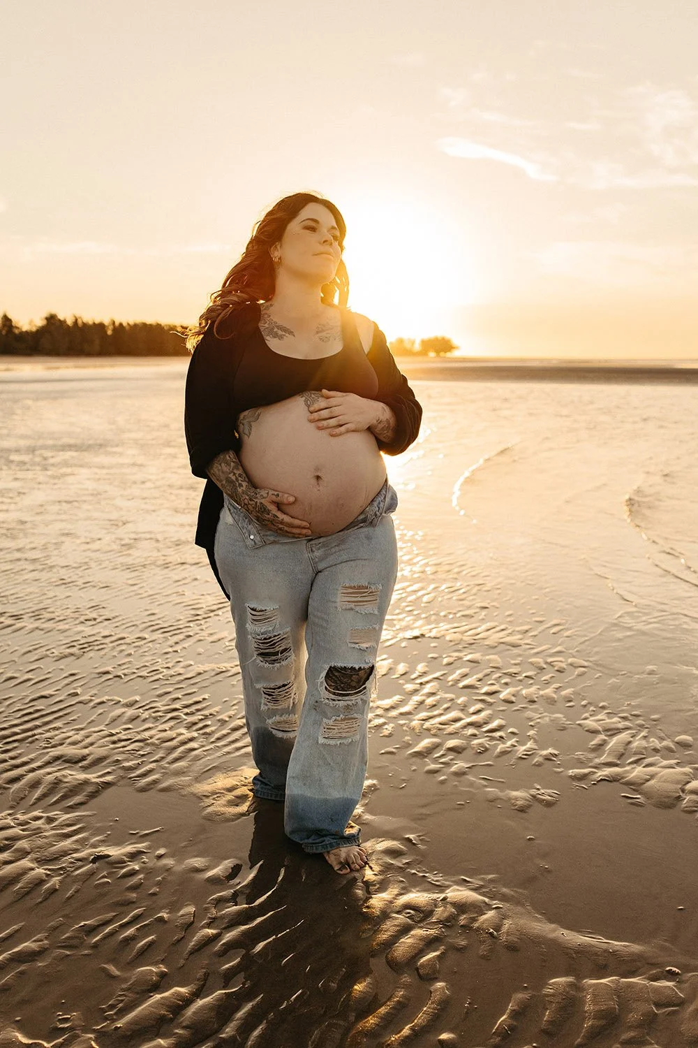 A pregnant woman wearing jeans on the beach at golden hour.