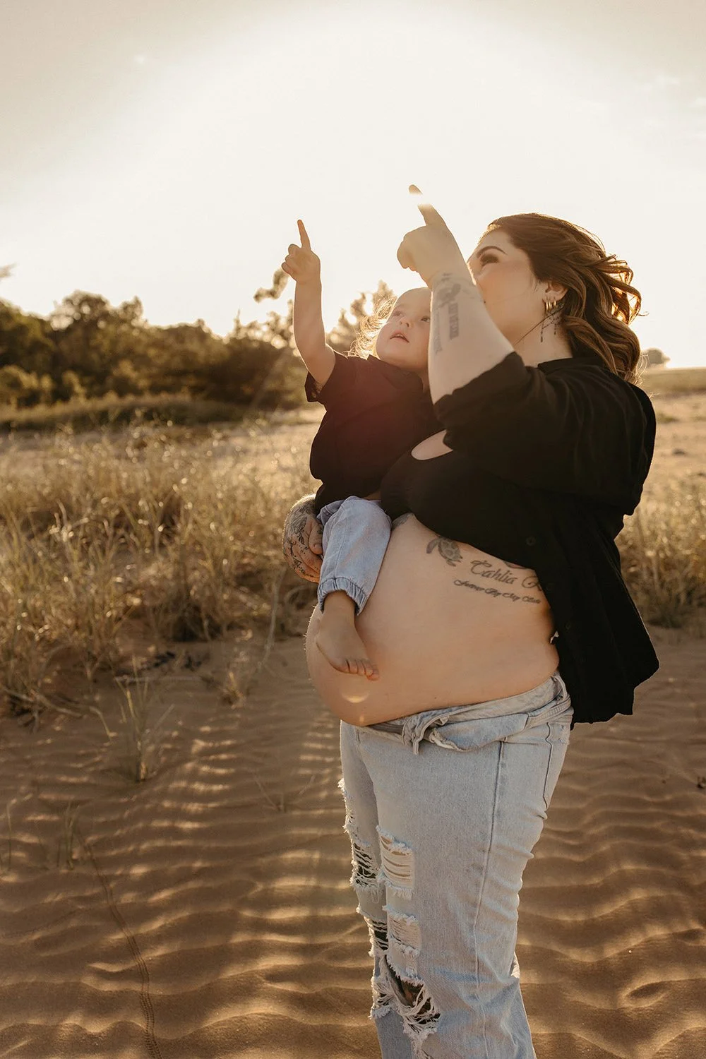 A pregnant woman holding her toddler, outside at golden hour. They point at the sky. Family photography session, Captured by Cody NT