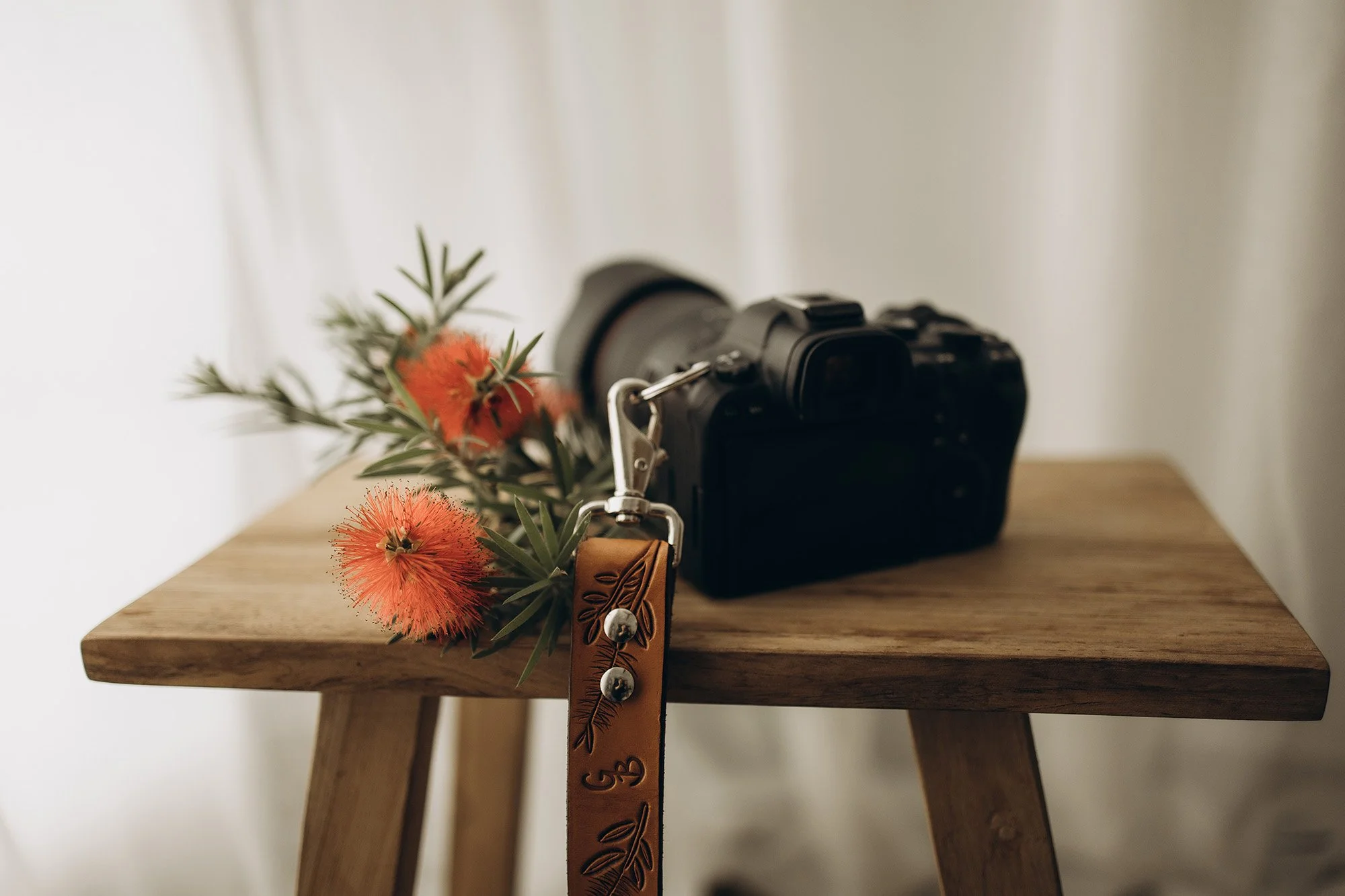 A professional photo of a camera with a leather strap on a timber stool with red bottlebrush flowers