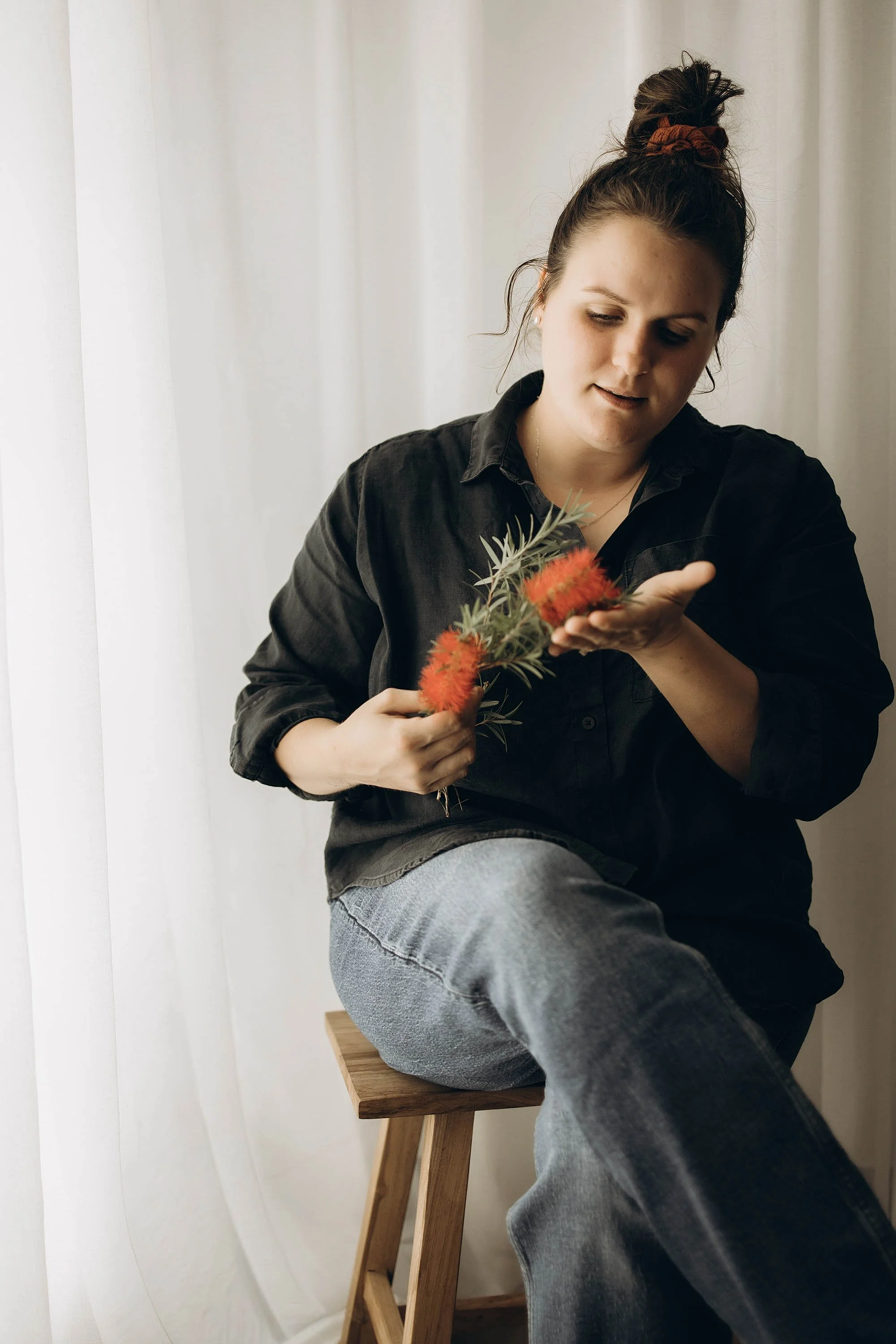 A photo of photographer Captured by Cody in her Darwin studio wearing a dark linen shirt, holding a red Australian flower