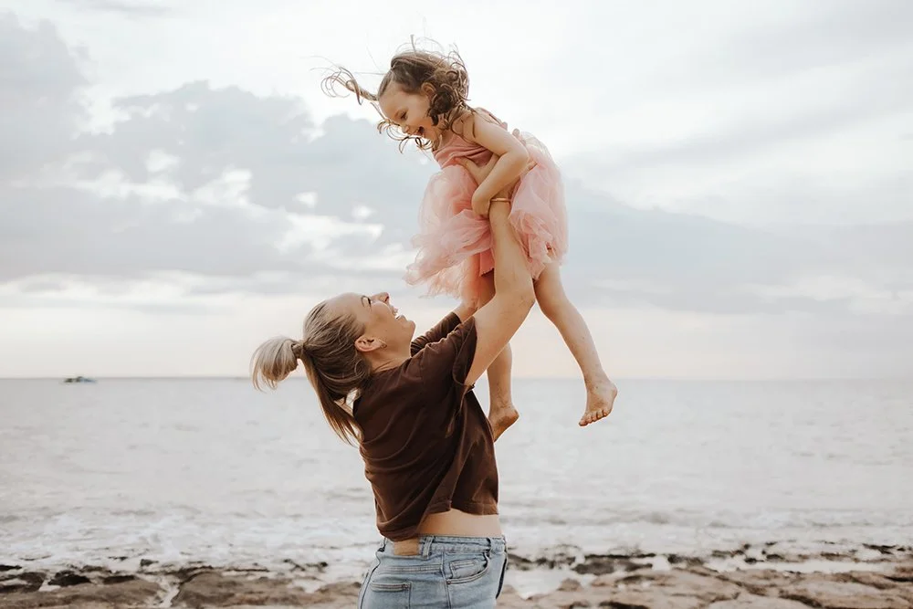 A mother lifting her young daughter into the air by the sea in NT by Captured by Cody family photographer.