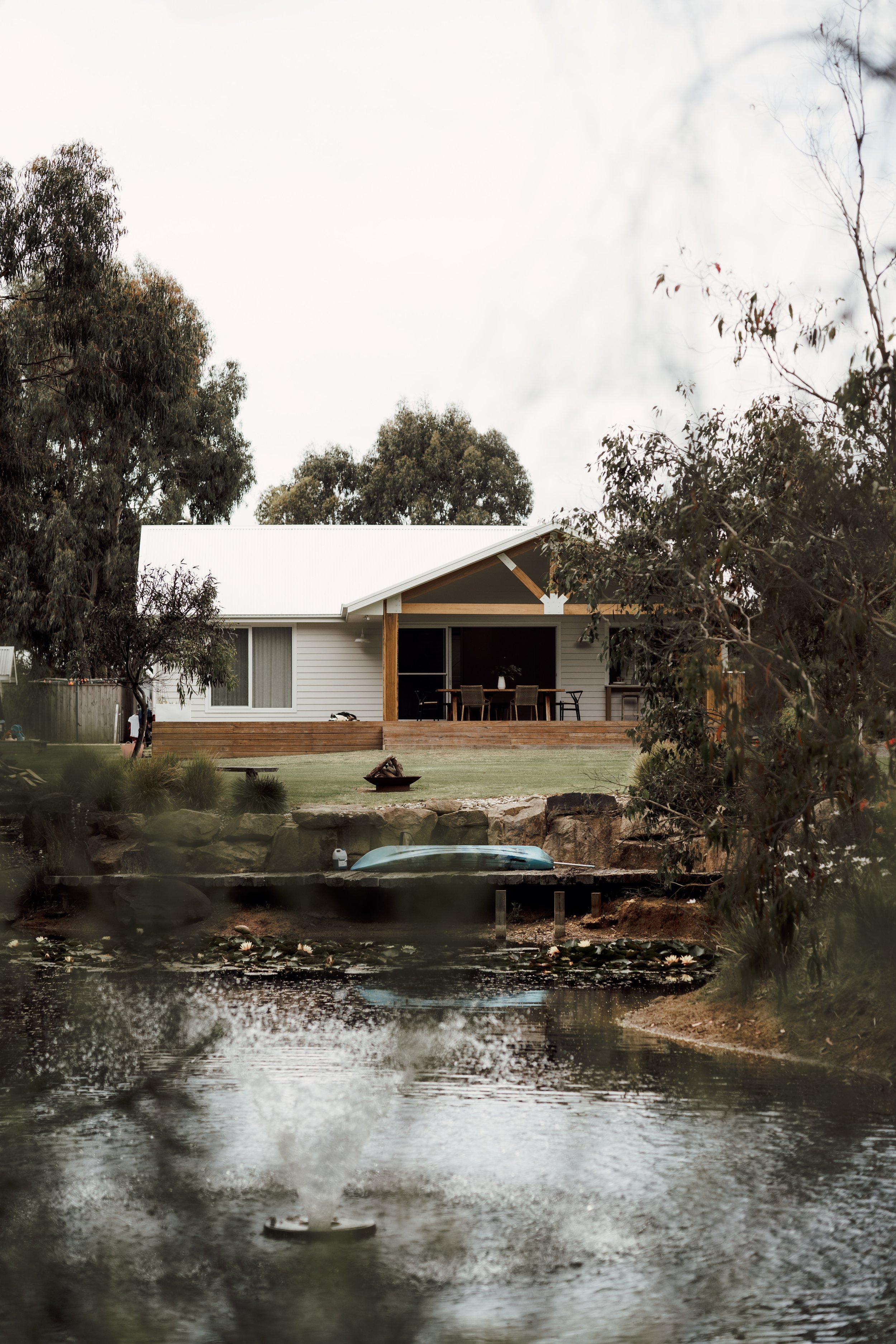A house with a veranda facing a pond with a boat and a fire pit, surrounded by trees under a cloudy sky.