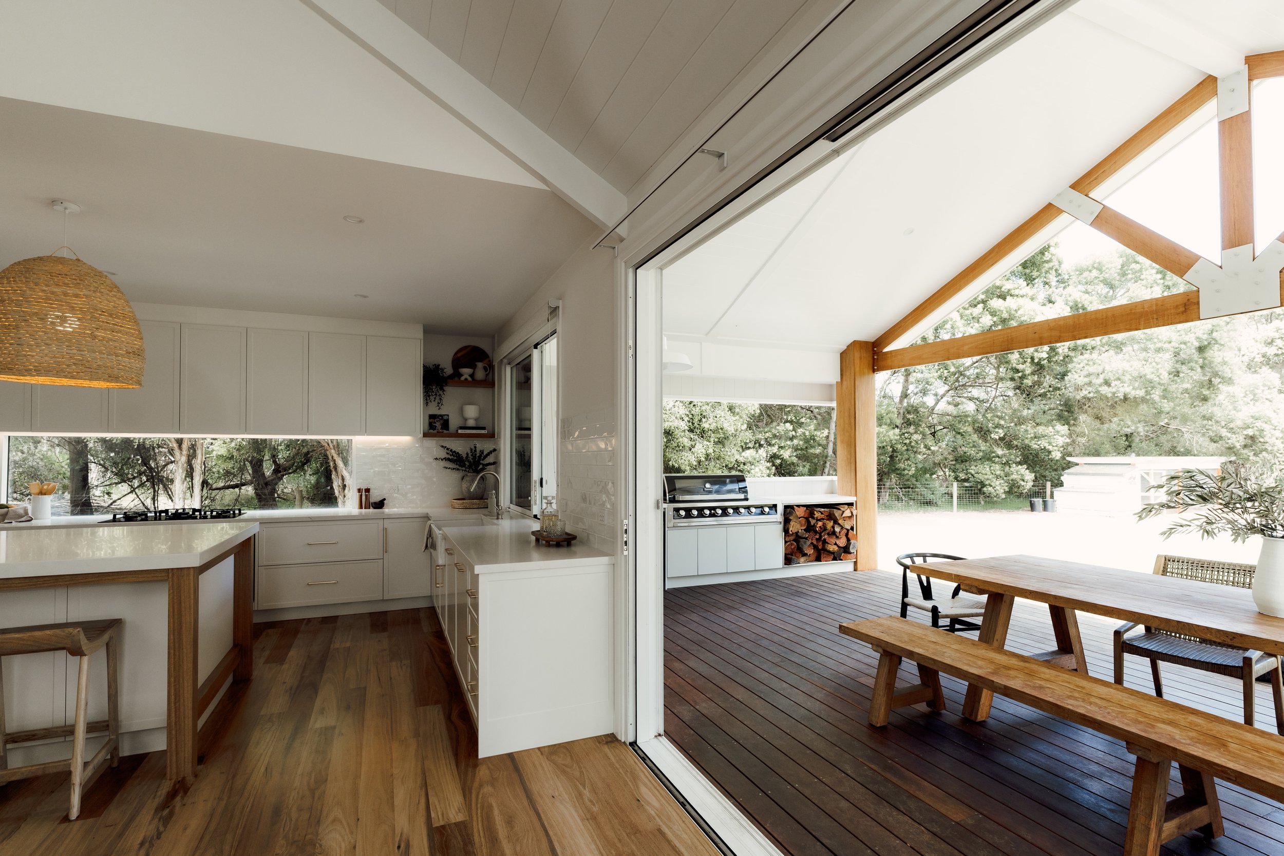 Kitchen with white cabinets, a large window, and an outdoor porch area with a wooden table and benches, surrounded by trees.