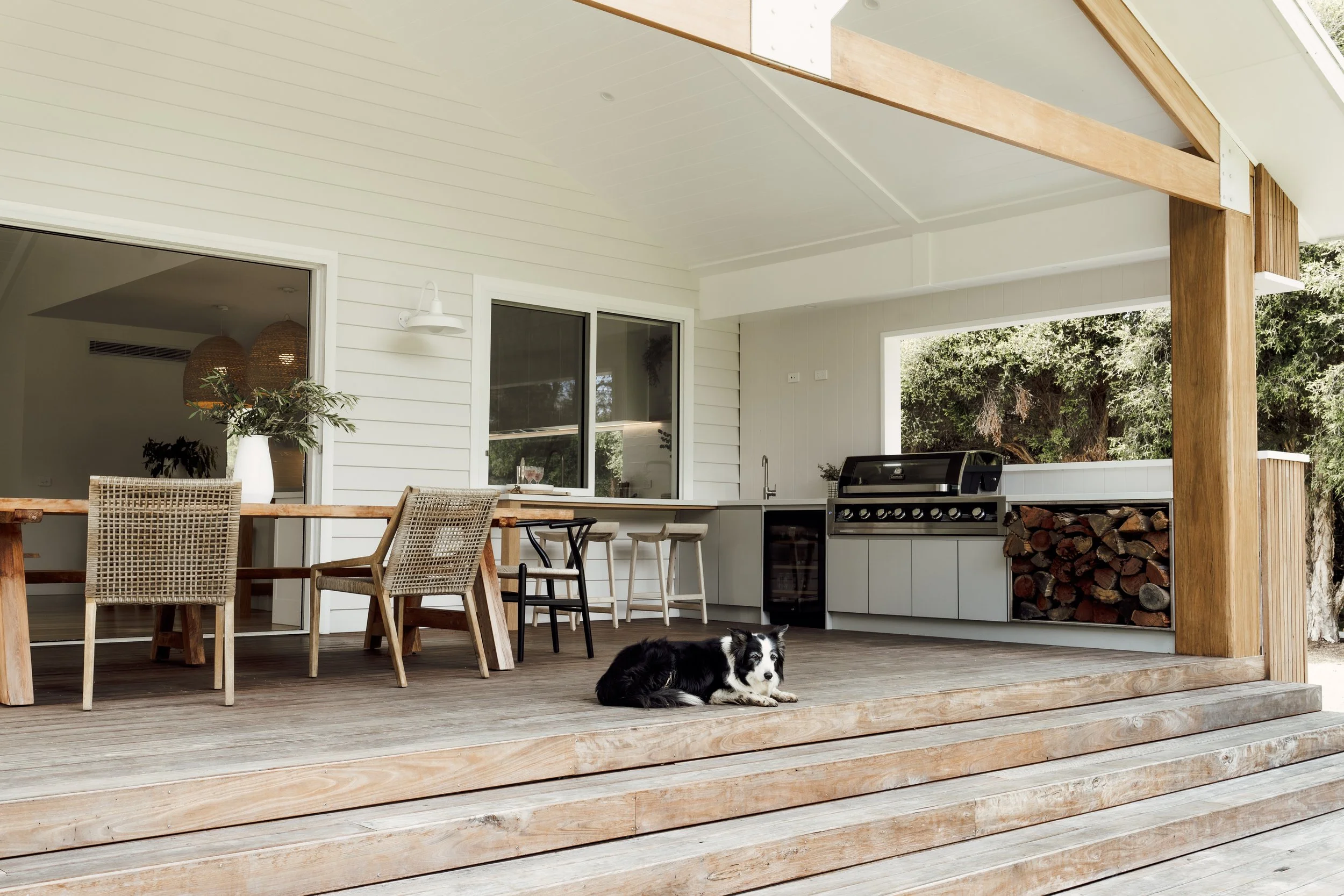 An outdoor porch with wooden flooring, white siding, and a built-in outdoor kitchen with a barbecue grill, stacked firewood, and bar stools. There is a potted plant on a wooden dining table, and a dog lying on the porch.