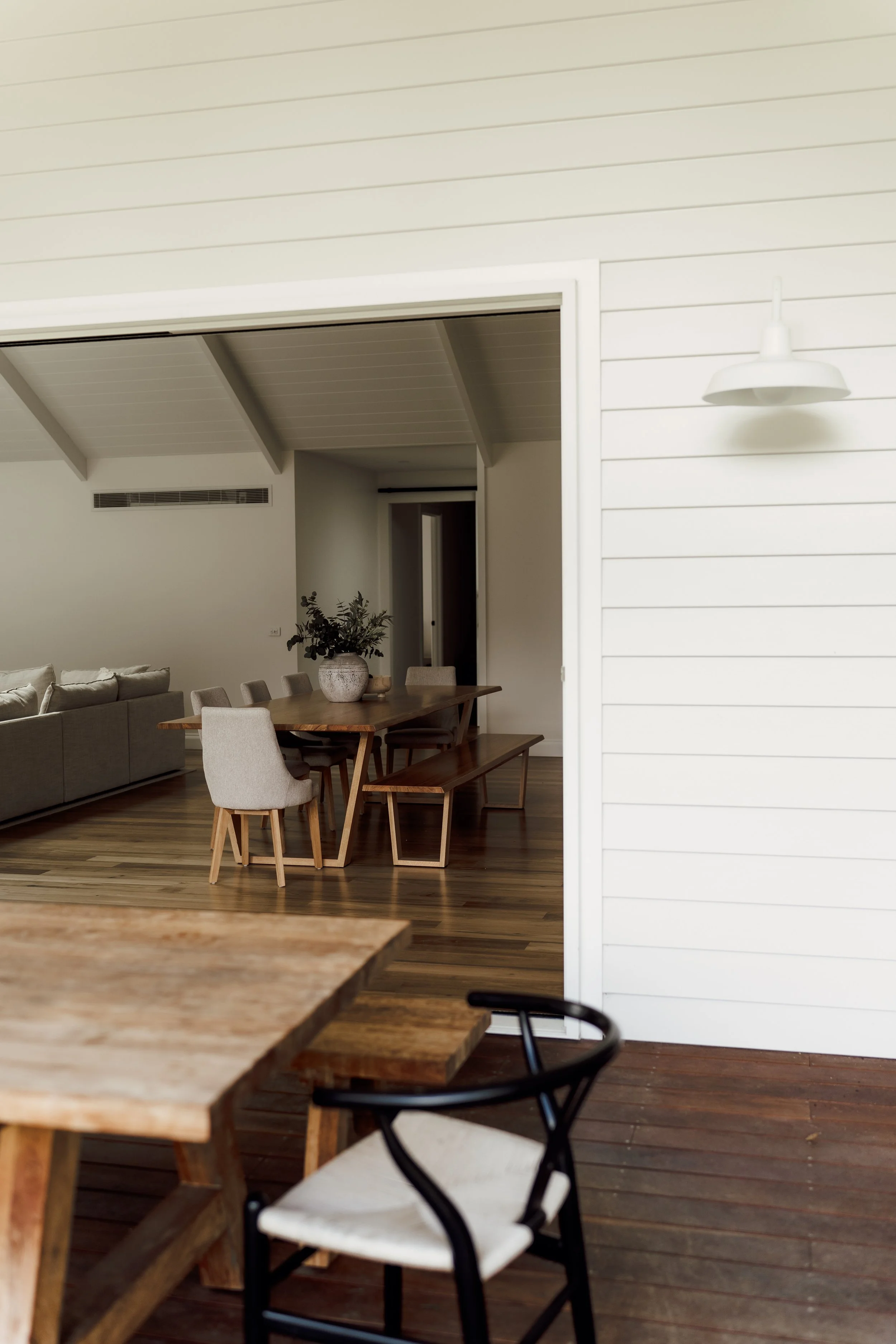 Open-concept dining and living room with wooden table, white chairs, gray sofa, indoor plant, ceiling light, and wooden flooring.
