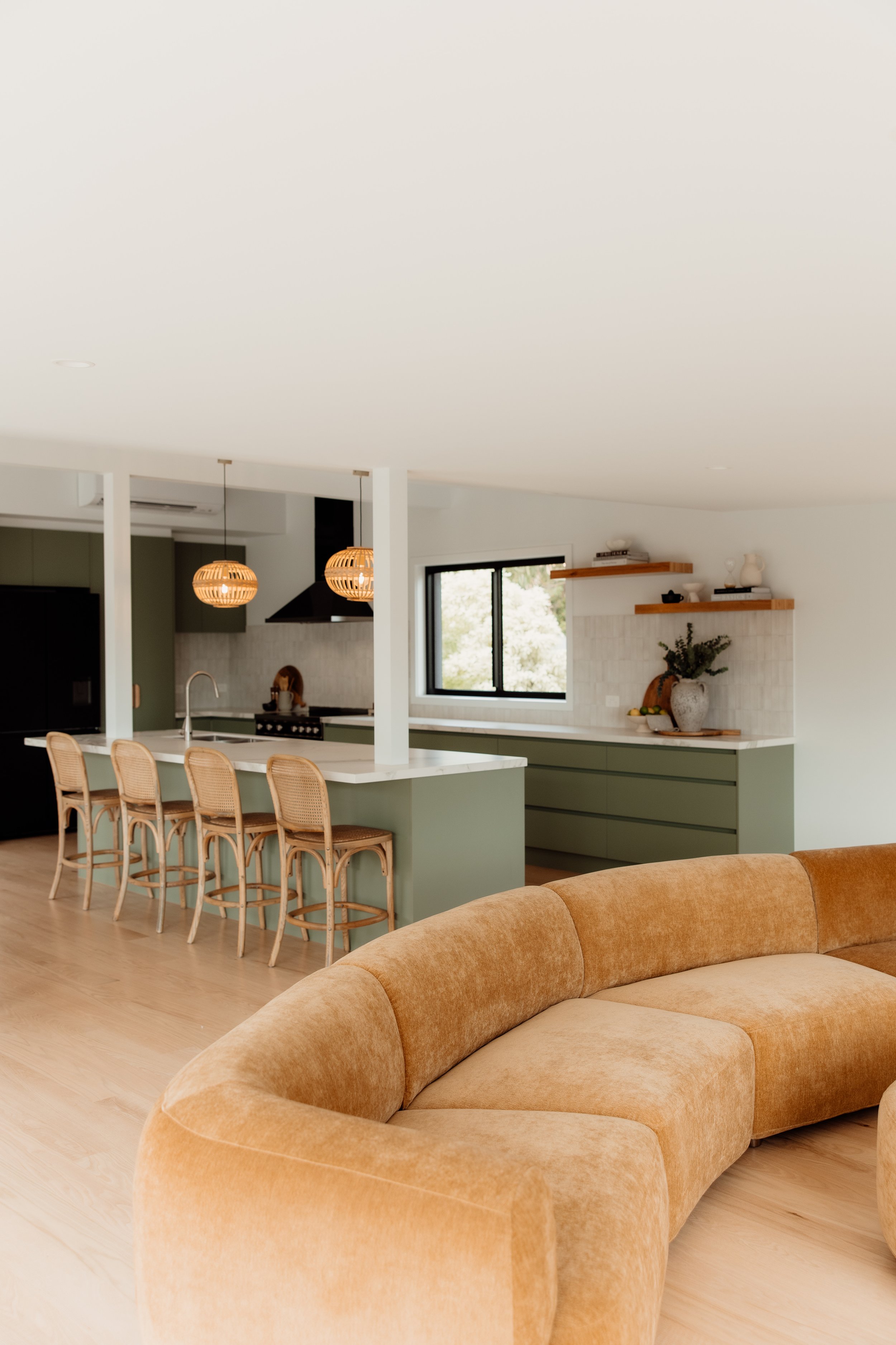 A modern open-concept kitchen and living area with a beige curved sofa, kitchen island with bar stools, hanging pendant lights, window with view outside, and shelves with decor.