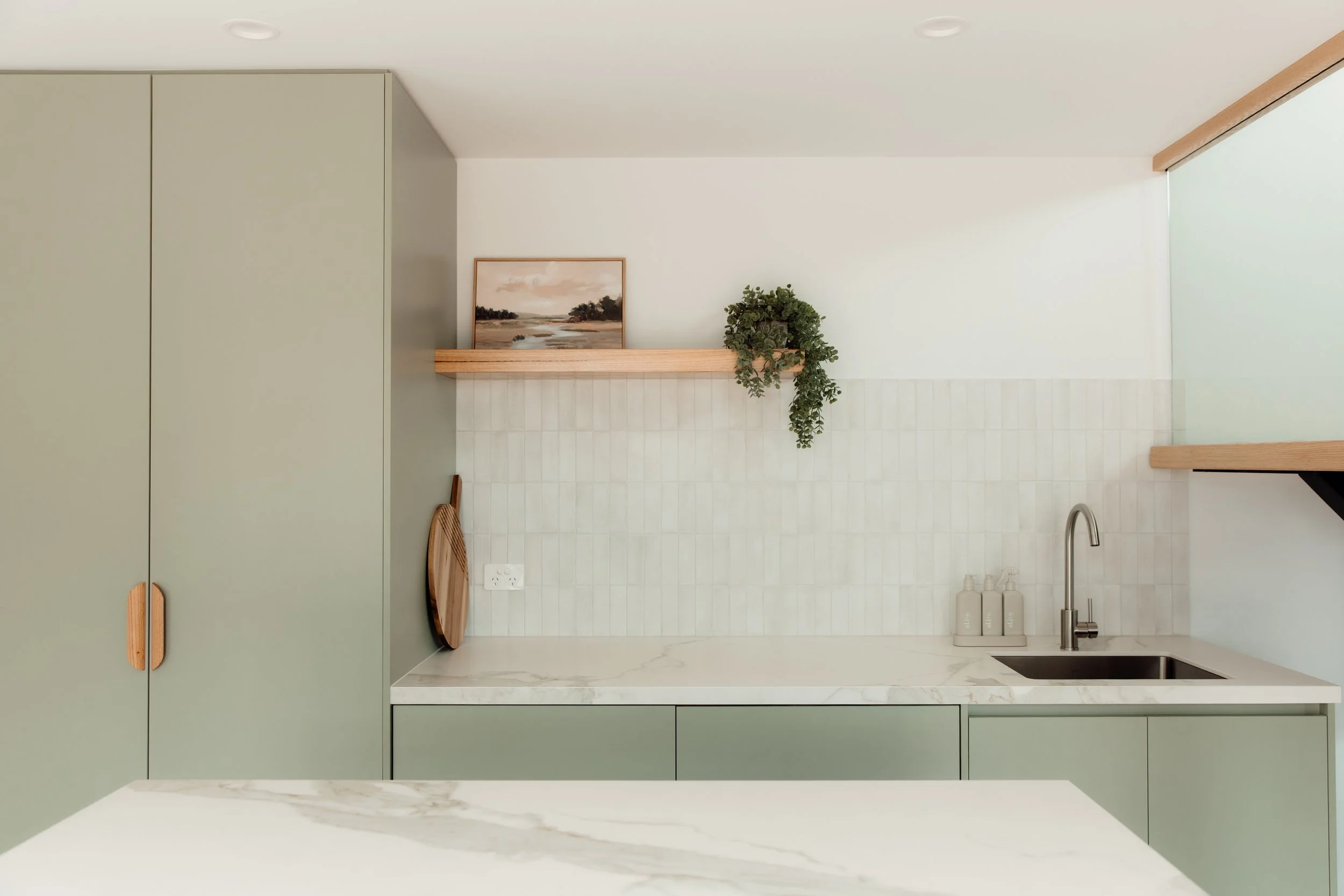 Minimalist kitchen with green cabinets, white marble countertop, white tile backsplash, a small sink with a faucet, three soap dispensers, a floating wooden shelf with a painting and a potted plant, and a wooden chair with rounded backrest.