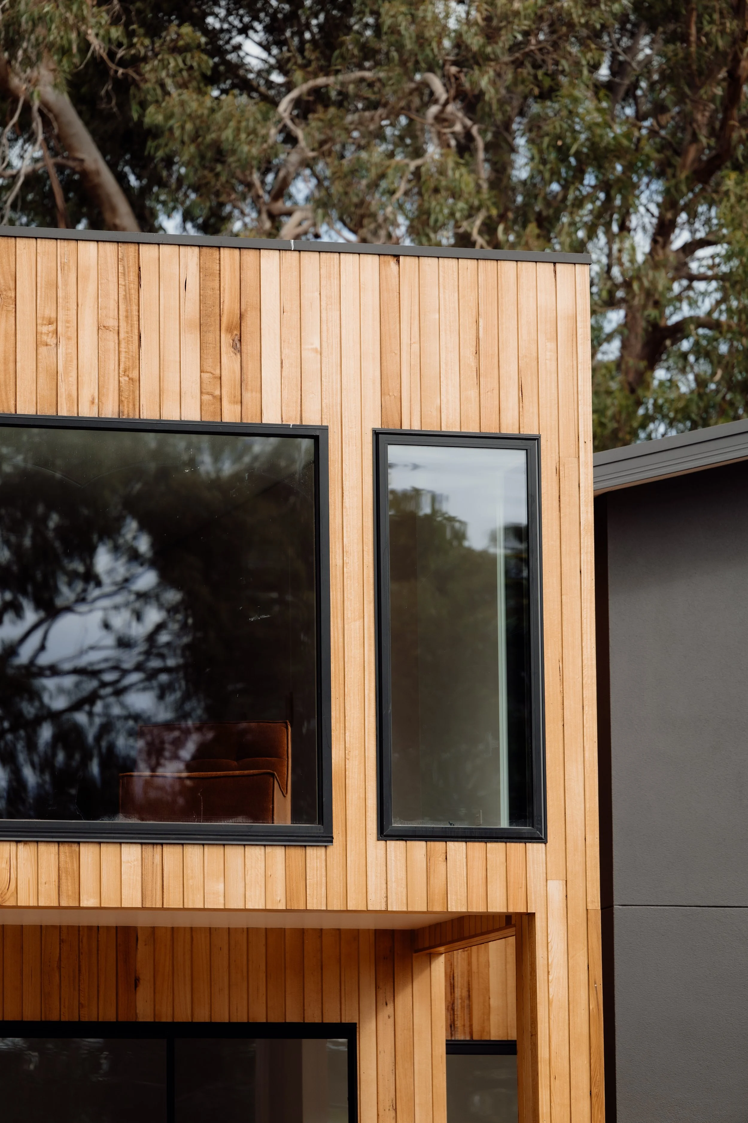 Close-up of a modern house exterior with vertical wooden siding, large black-framed windows, and a tree in the background.