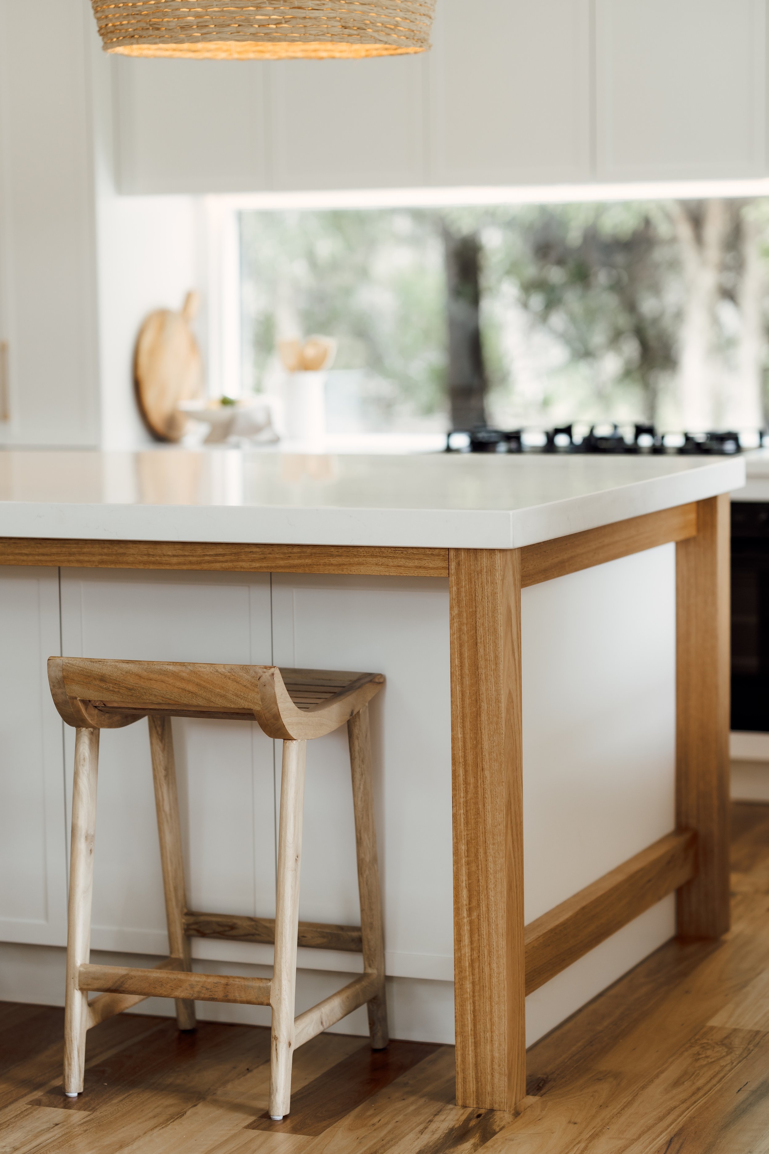 Close-up of a kitchen island with a wooden frame and a white countertop, with wooden bar stool underneath near a window with trees outside.