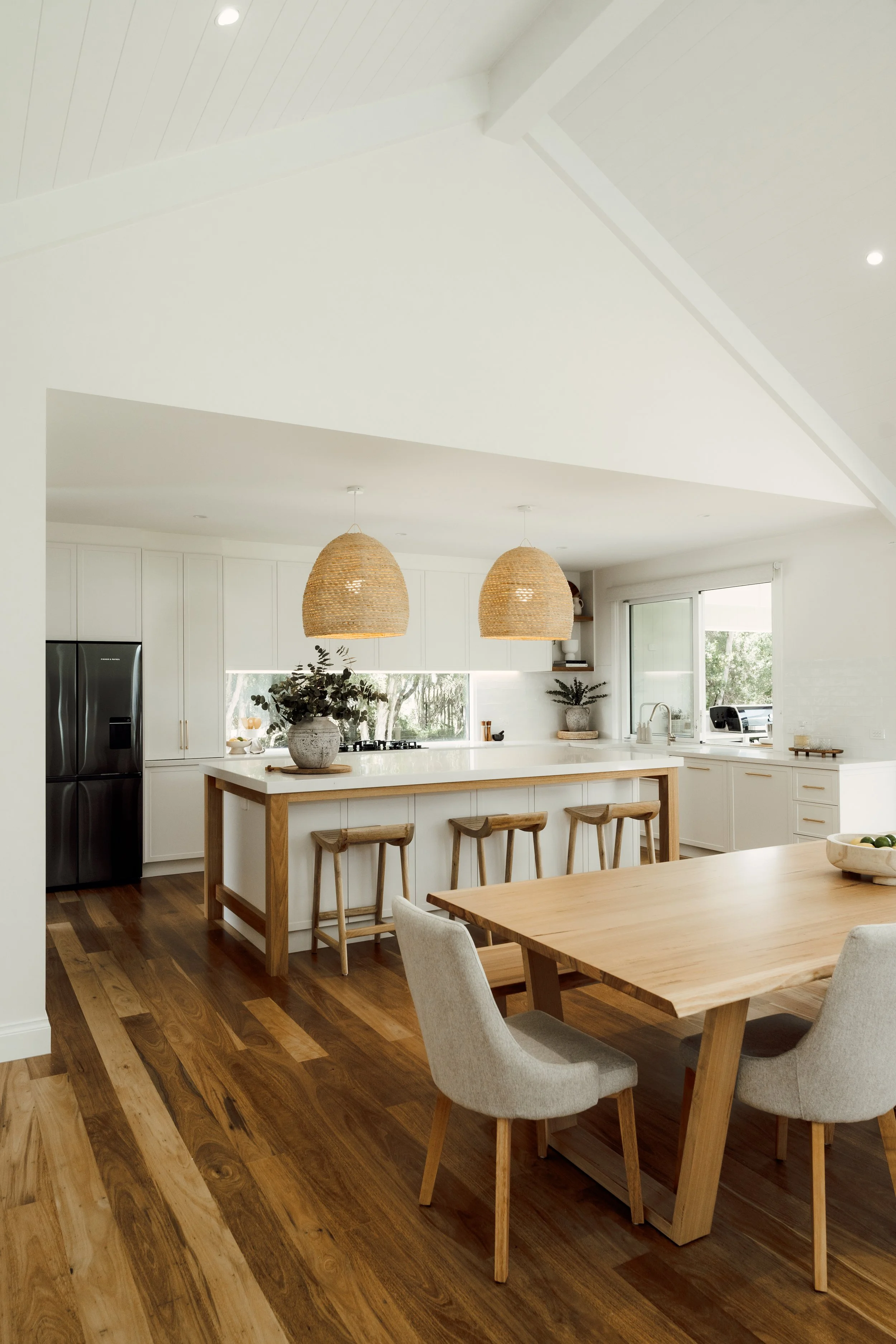 A modern kitchen with white cabinets, a large island with a wooden frame, and barstools. Two hanging rattan pendant lights, a potted plant on the island, and a window with a view of trees outside.