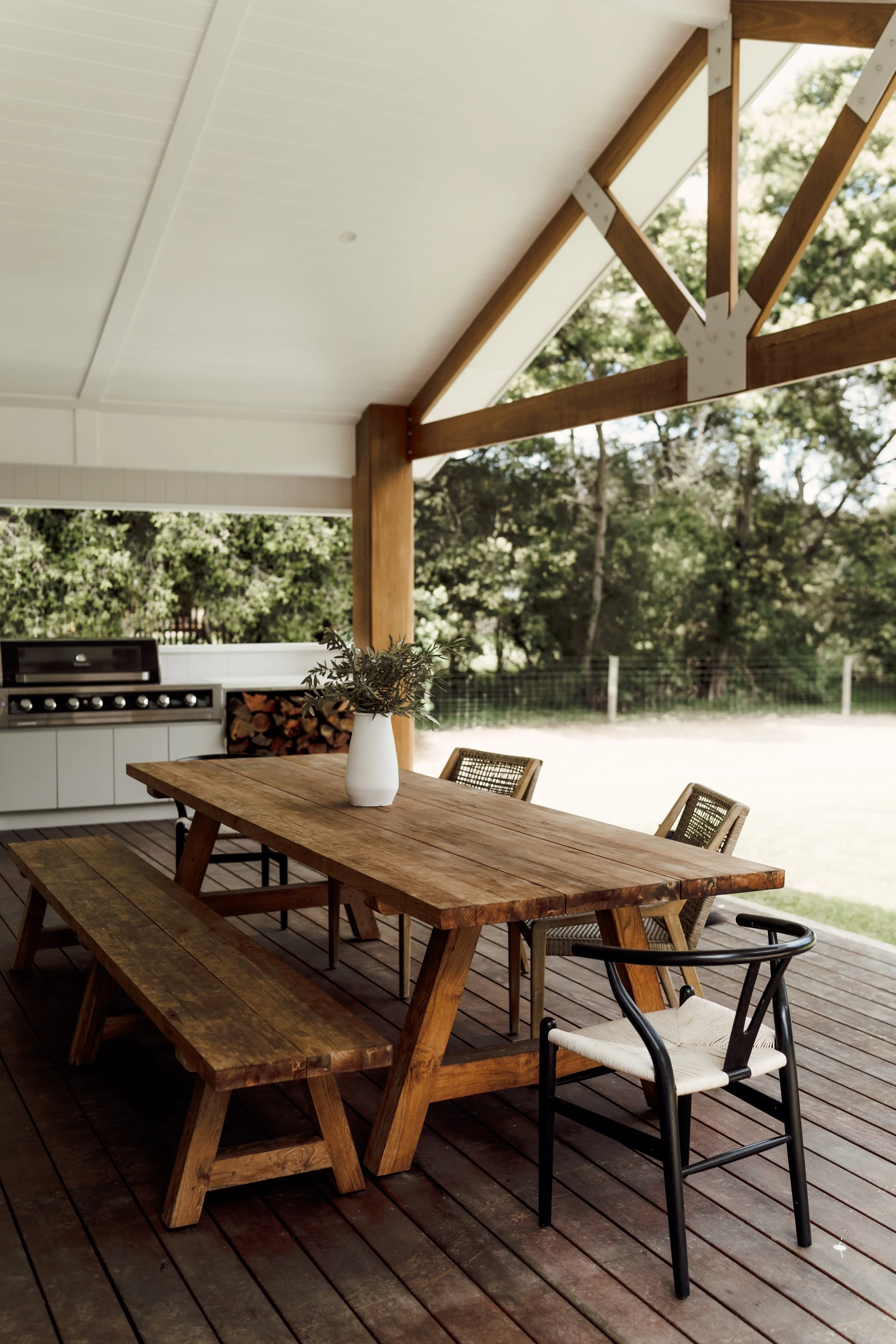 An outdoor patio with a large rustic wooden dining table, a bench on one side, and various chairs including a black metal chair and a rattan chair. A white vase with greenery decorates the table. The patio has a wooden deck floor and is covered with 