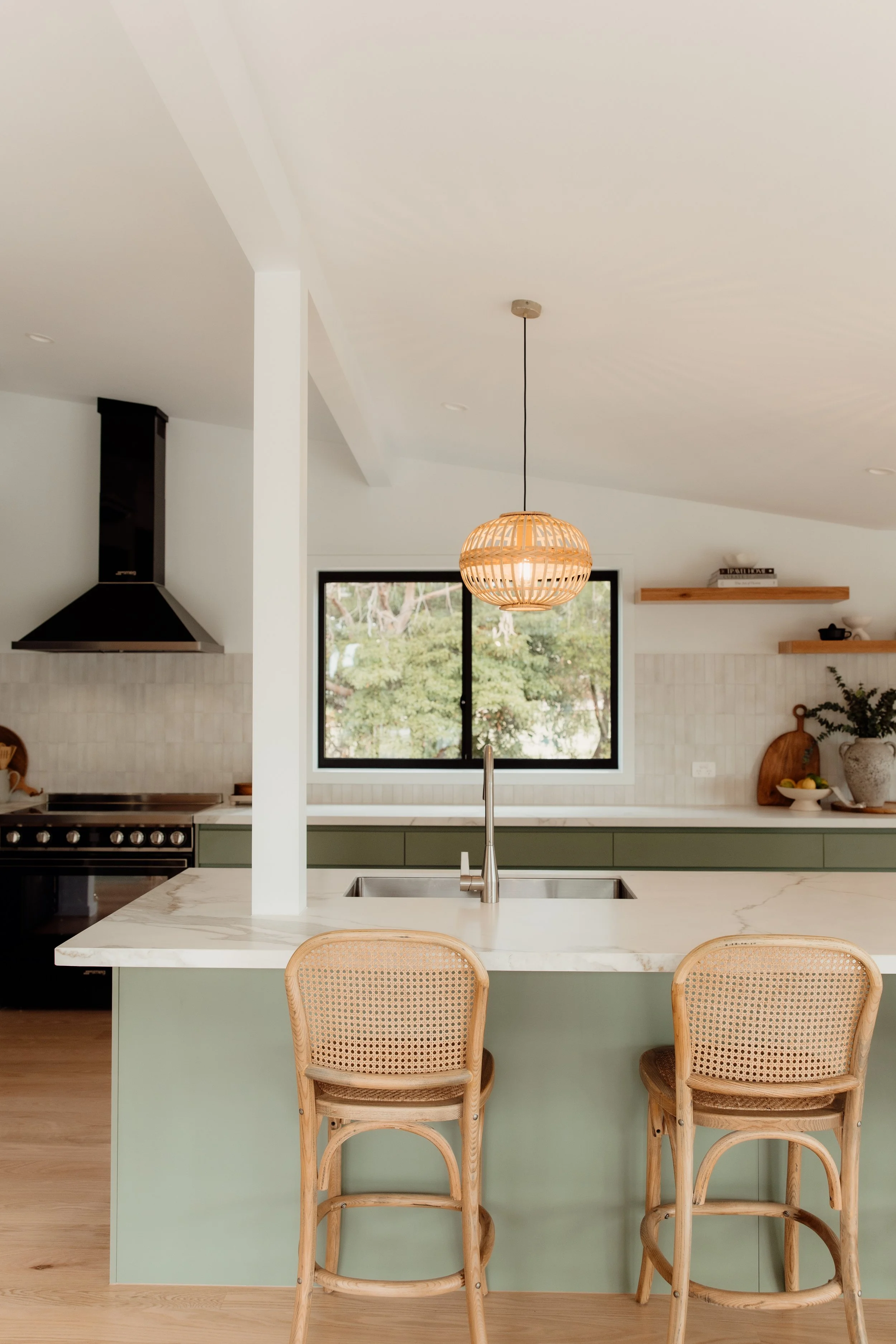 Modern kitchen with a white marble island, green lower cabinets, a hanging woven pendant light, two wicker chairs at the island, a window showing greenery outside, and open wooden shelves with decor and kitchen items.
