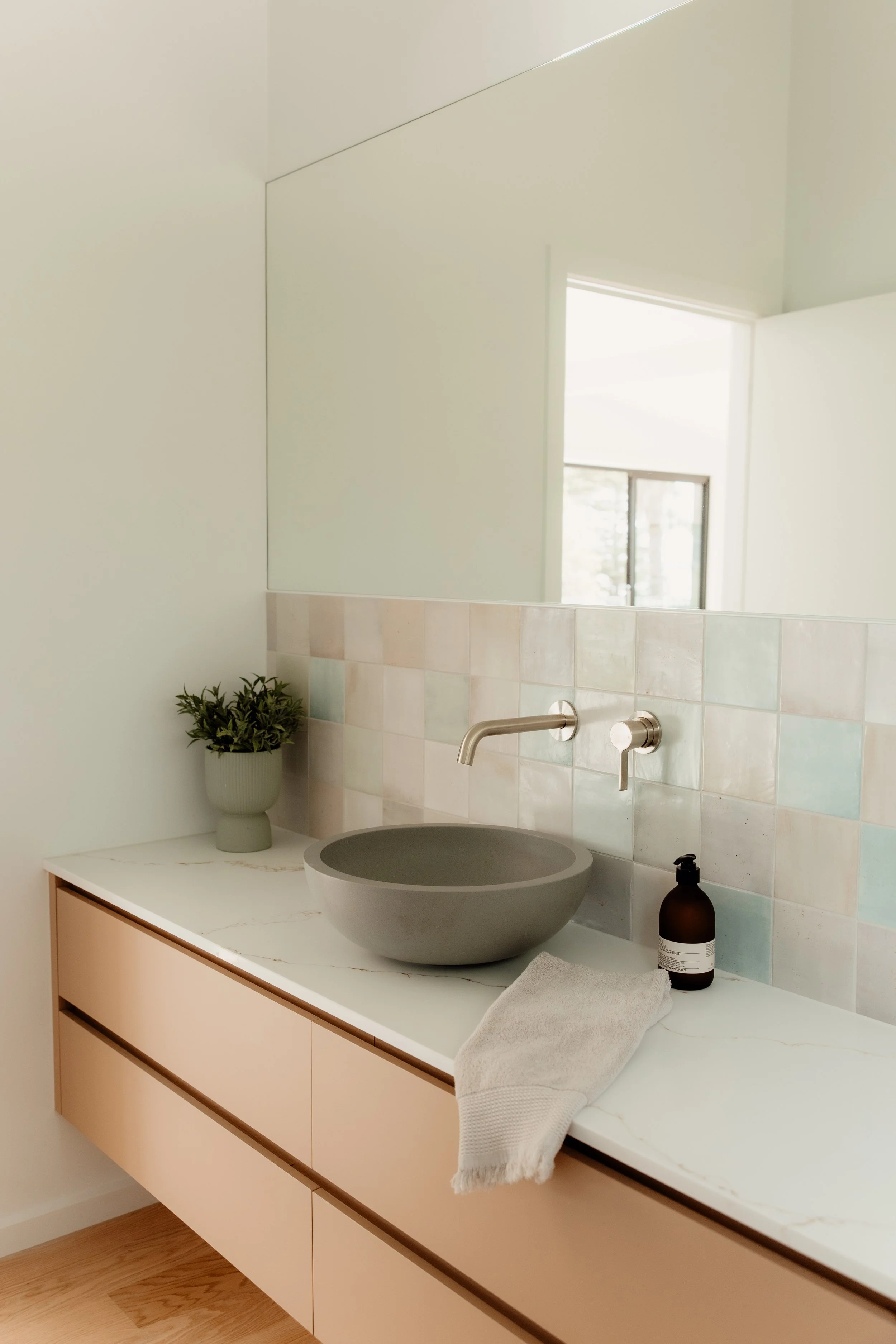 Modern bathroom with a large mirror, a floating vanity with drawers, a round vessel sink, a wall-mounted faucet, a brown soap dispenser, a towel, a potted plant, and a window reflected in the mirror.