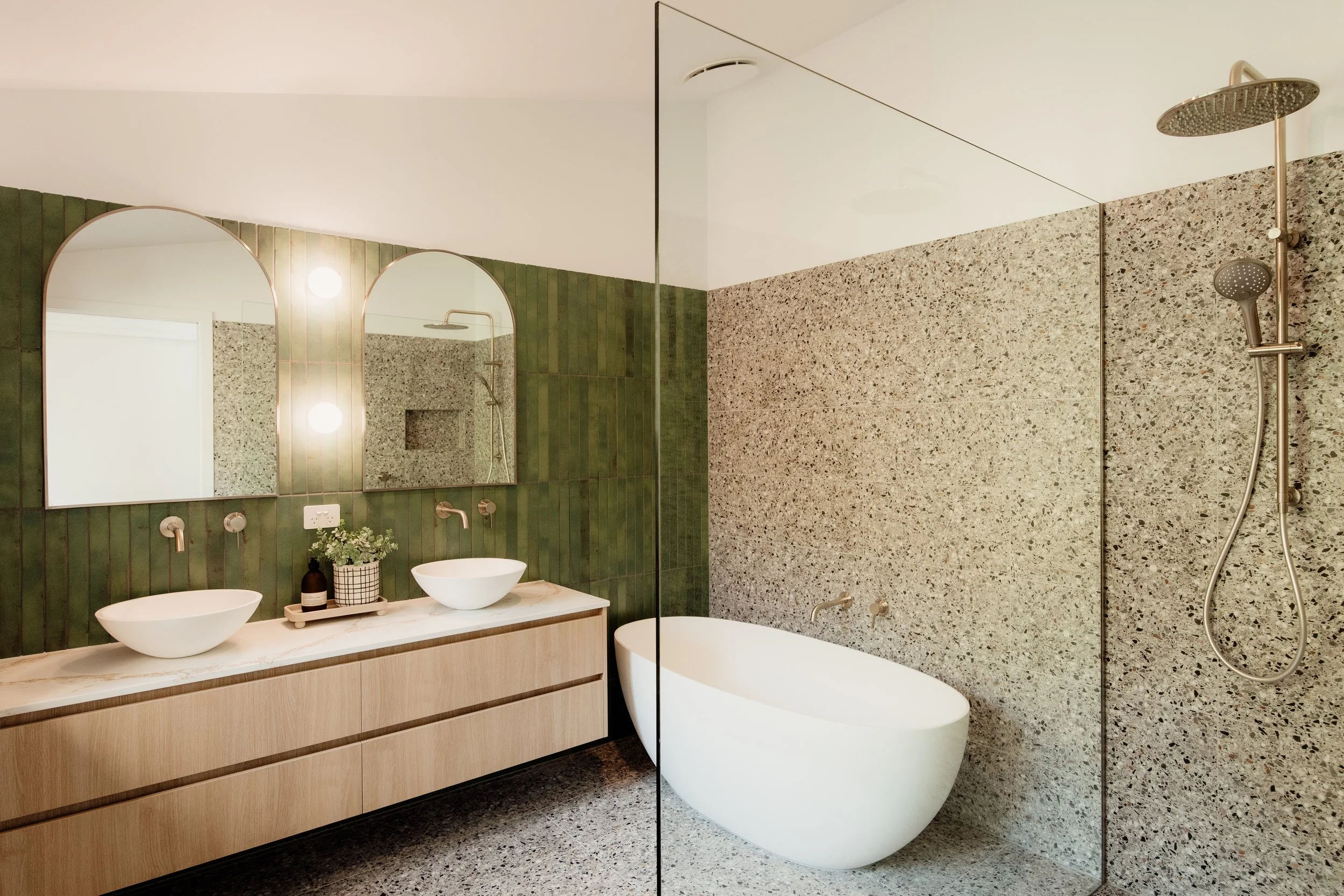 Modern bathroom with a double sink vanity, round vessel sinks, green tiled wall, large mirror, and a shower area with pebble stone walls, a rainfall showerhead, and a handheld shower.