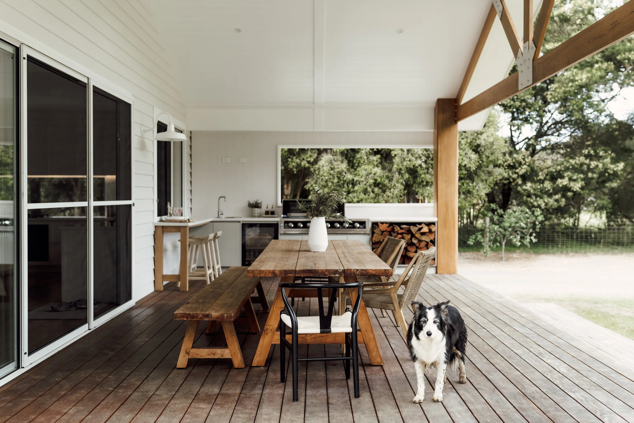 A covered outdoor patio with a wooden dining table, chairs, and a black and white dog standing on the wooden deck, with a background of green trees.