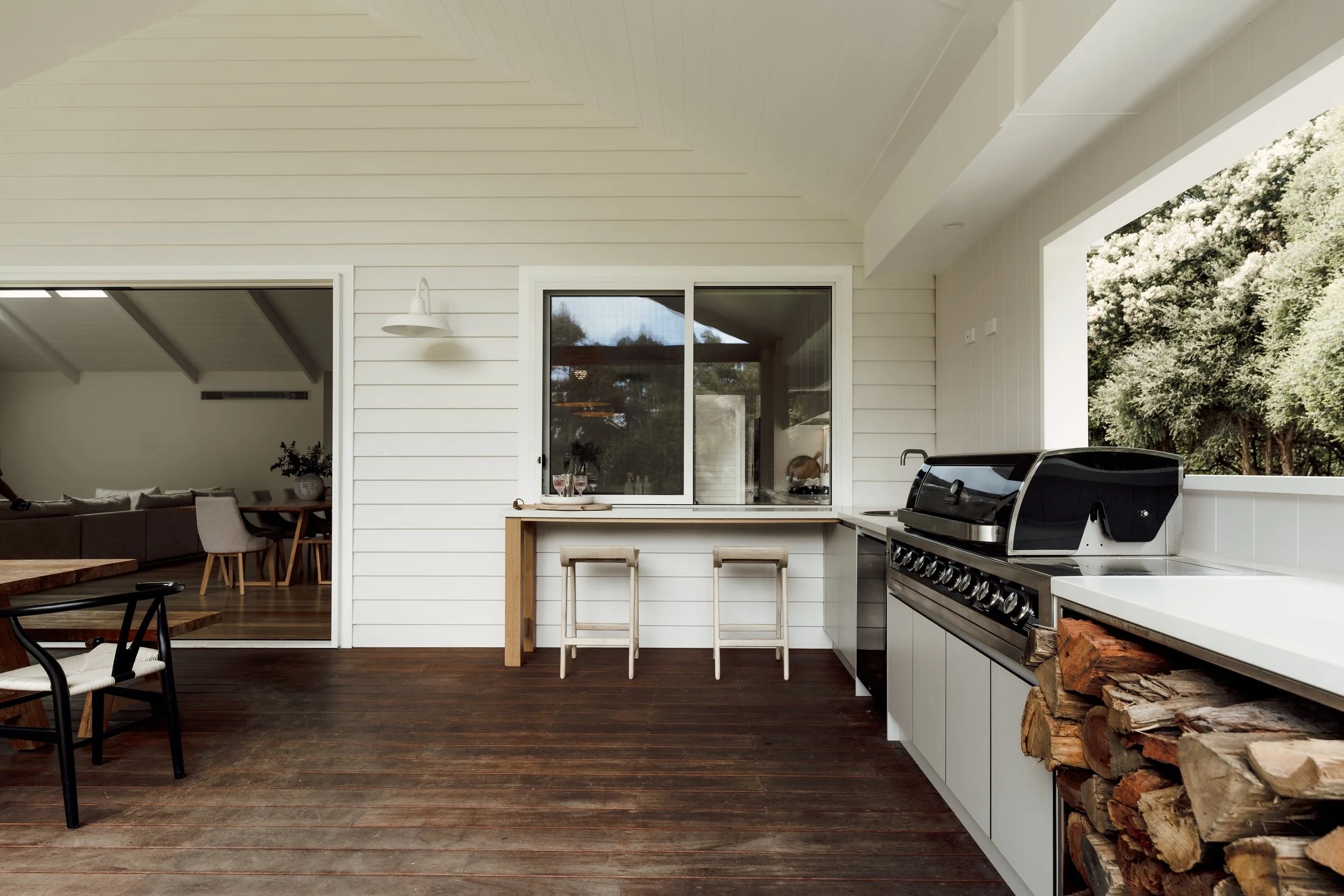 Outdoor kitchen with a black grill, stacked firewood, and a white countertop under a large open ceiling with a window and sliding glass door leading into interior dining area.