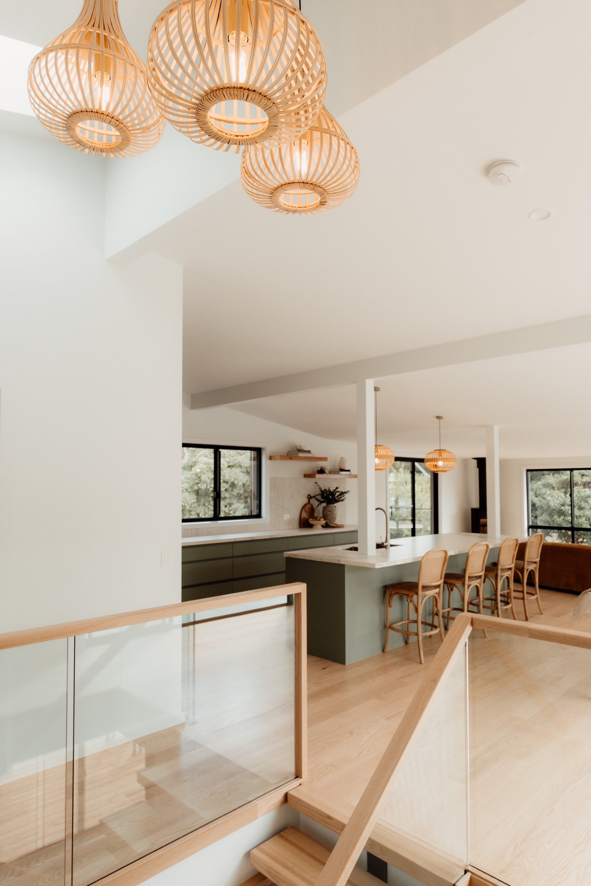 Modern kitchen with a white island, green lower cabinets, and woven pendant lights hanging from the ceiling. Large windows let in natural light, and a wooden staircase is visible in the foreground.
