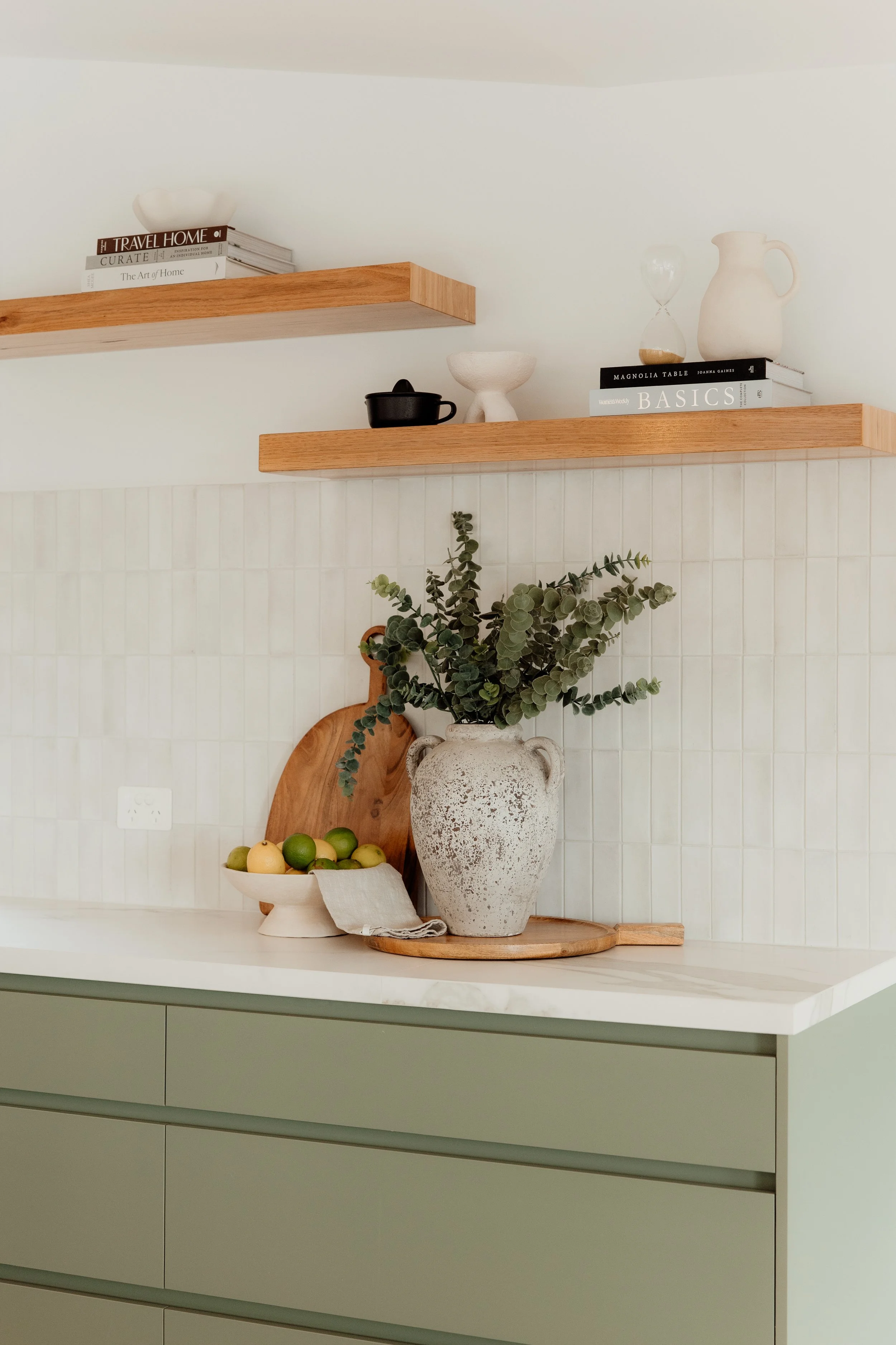 A modern kitchen shelf decor with books, a white vase, a black pot, an hourglass, a white bowl, and a large textured vase with eucalyptus leaves. A bowl of lemons and limes on the counter below.