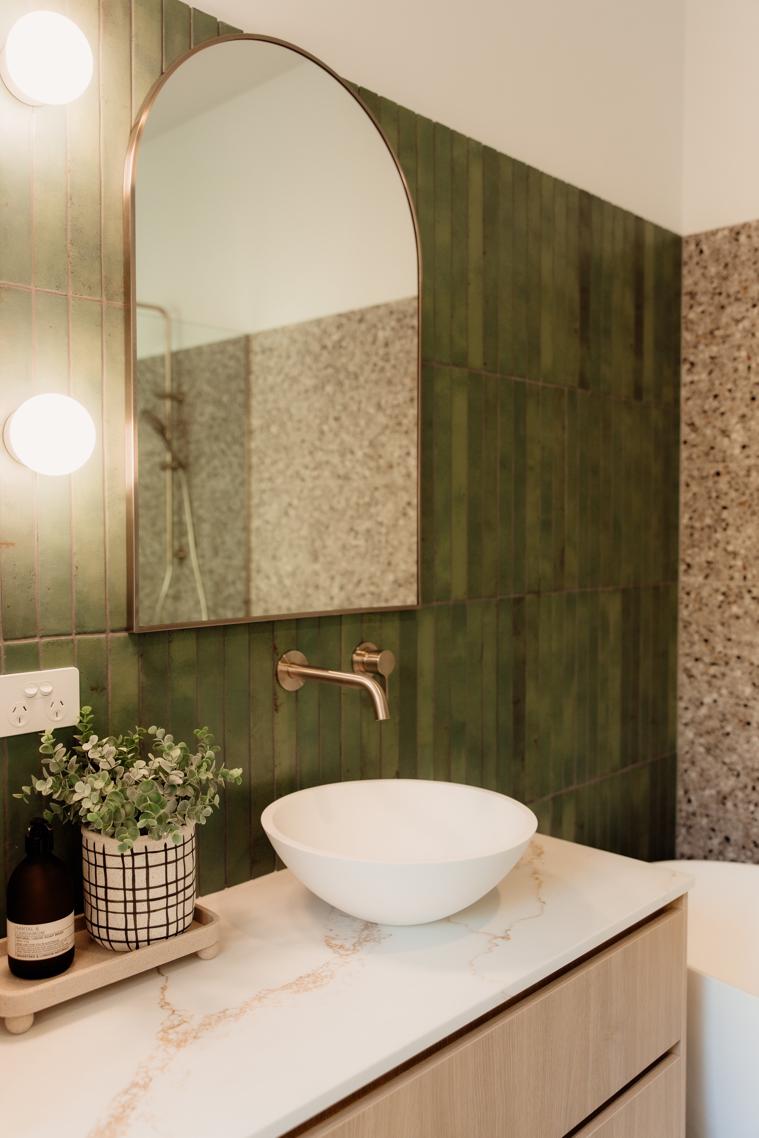 Bathroom vanity with a round white vessel sink on a marble countertop, a potted plant, a soap bottle, a large mirror, and green tiled walls.