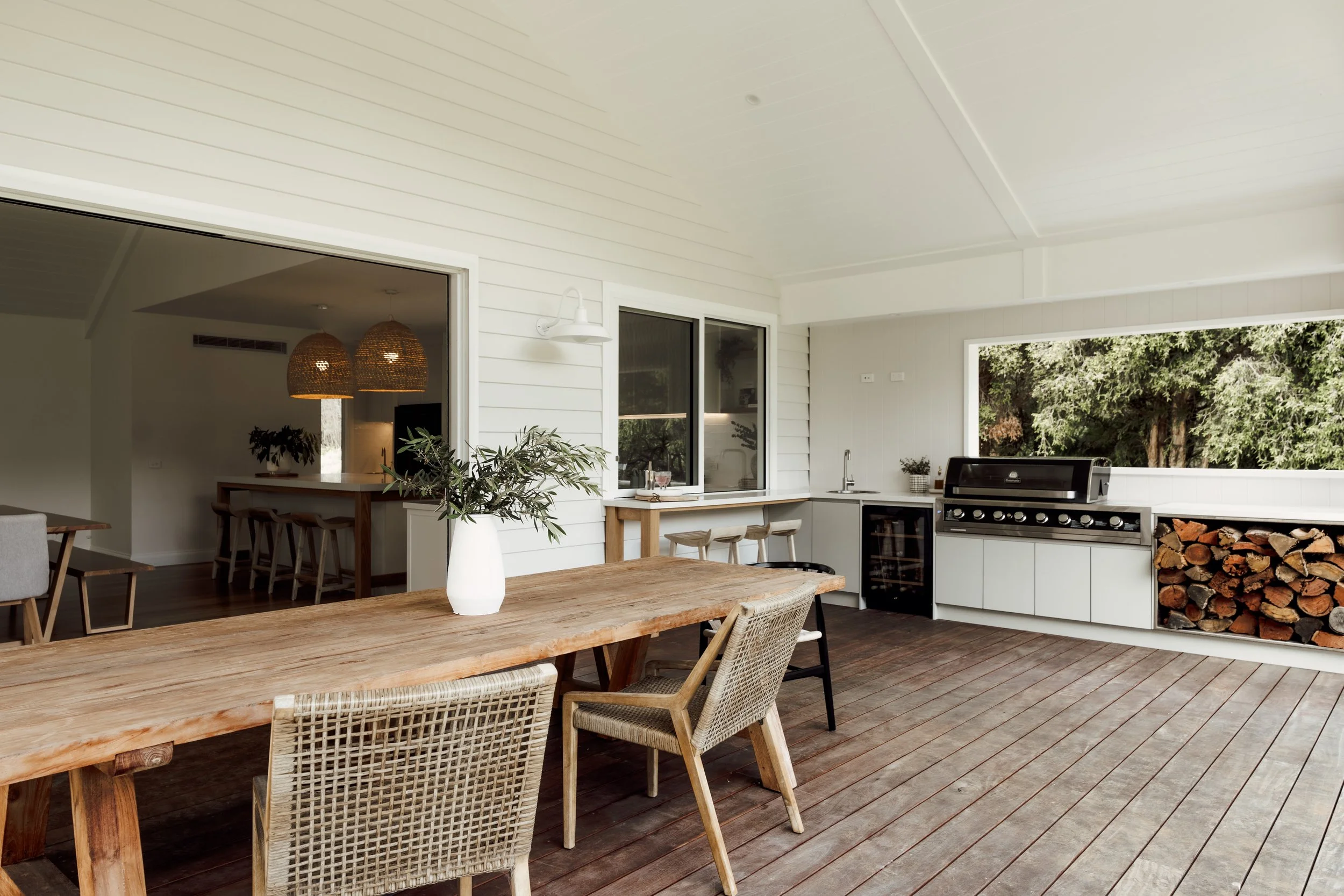 A spacious outdoor kitchen and dining area with white walls, a large wooden dining table, woven chairs, a gas grill, and a wood storage area, surrounded by green trees.