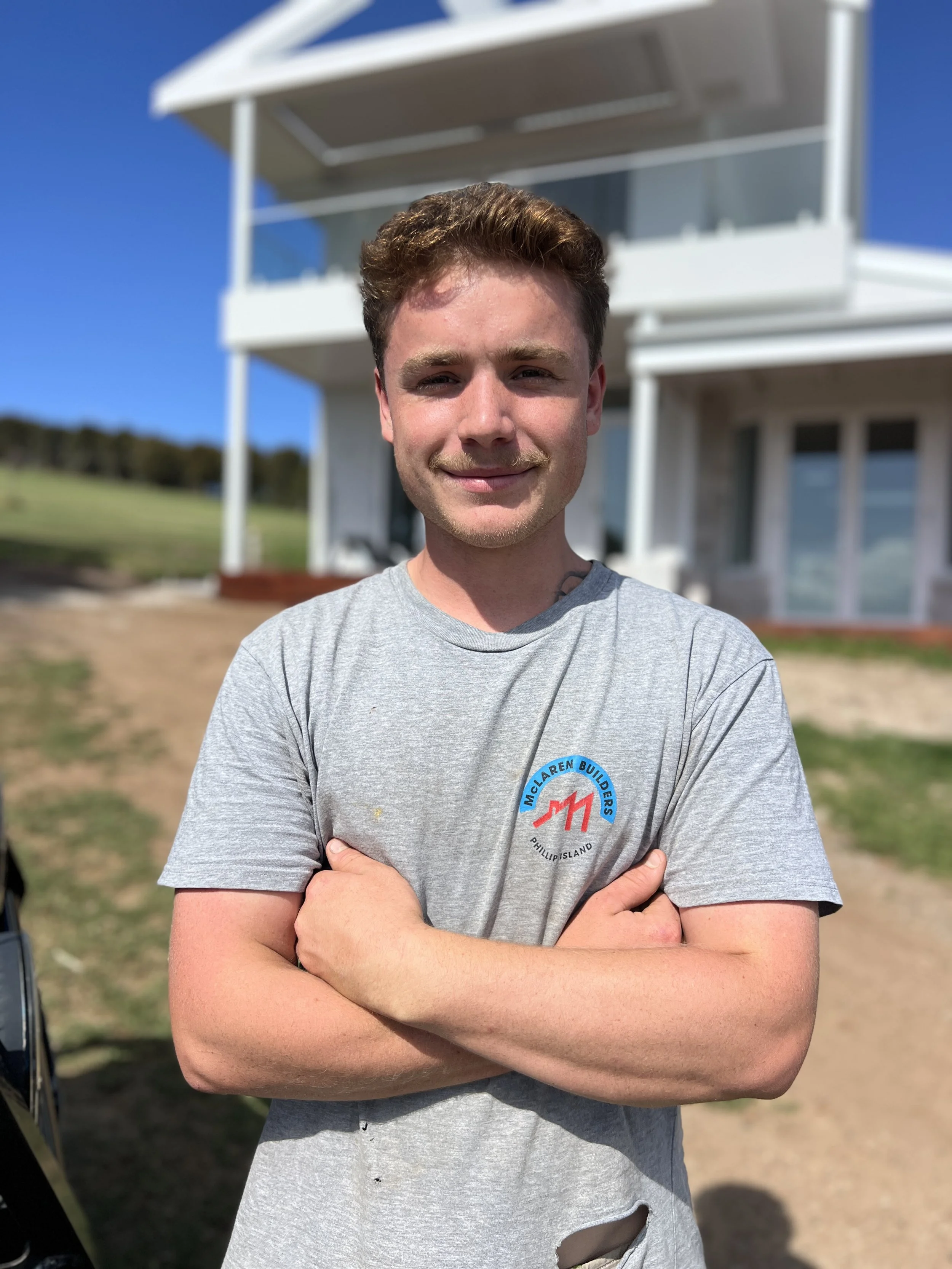 A young man with curly brown hair and a light complexion smiling outdoors with crossed arms, wearing a grey T-shirt with a logo that reads "McLaren Builders Philippi, WV". Behind him is a modern white building with multiple stories and a clear blue sky.