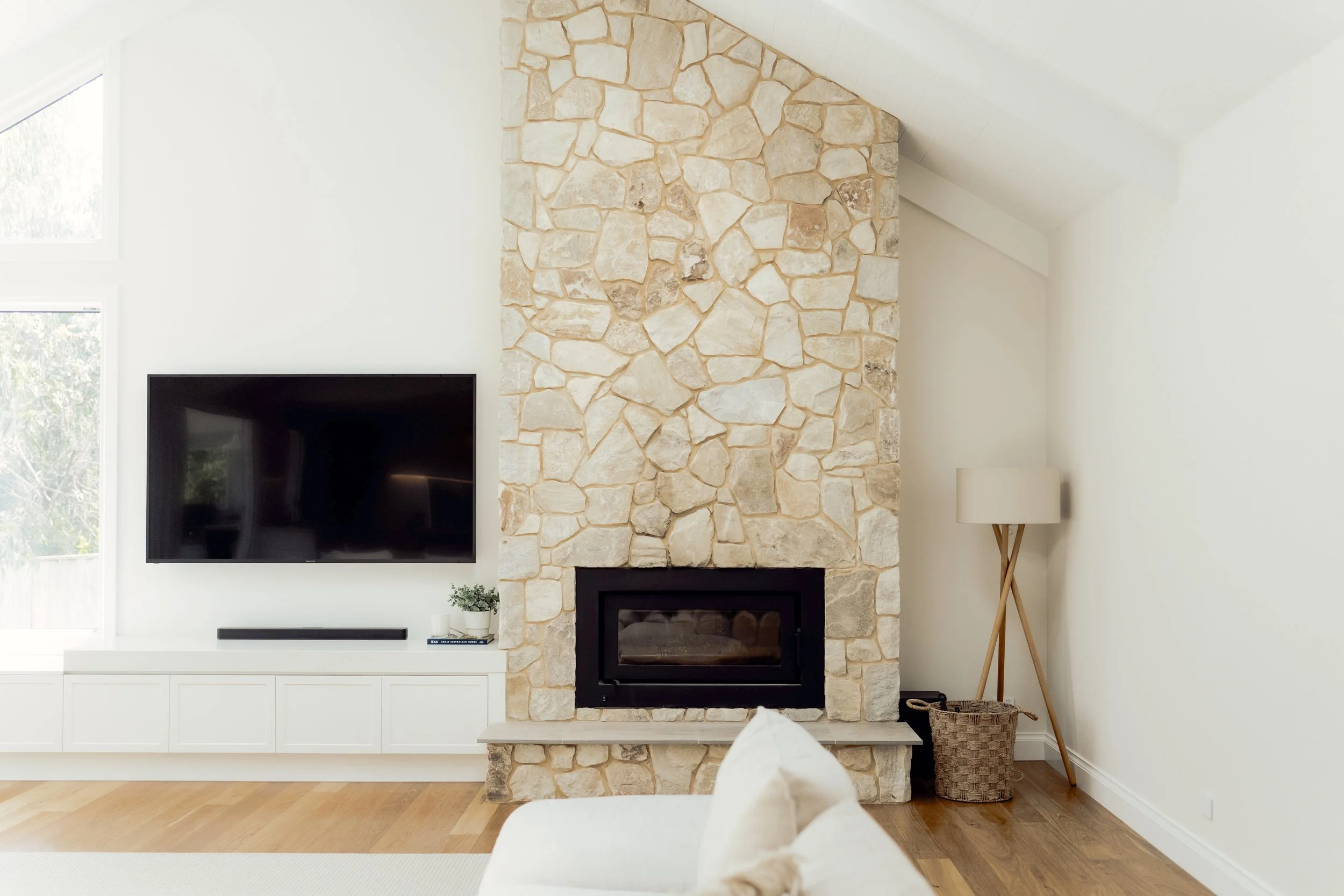A living room with a stone fireplace, flat-screen TV on the white wall, a window to the left, a white cabinet underneath the TV, a small potted plant and a speaker on the side, and a floor lamp with a wicker basket nearby.