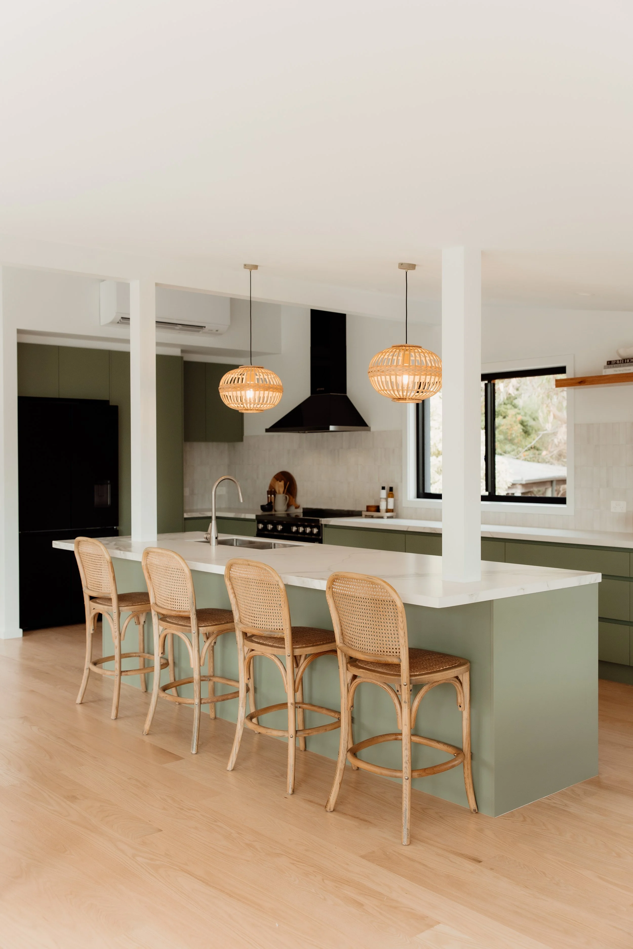 A modern kitchen with a white island counter, four rattan barstools, pendant lights, a black range hood, black refrigerator, green cabinets, a window, and wooden flooring.