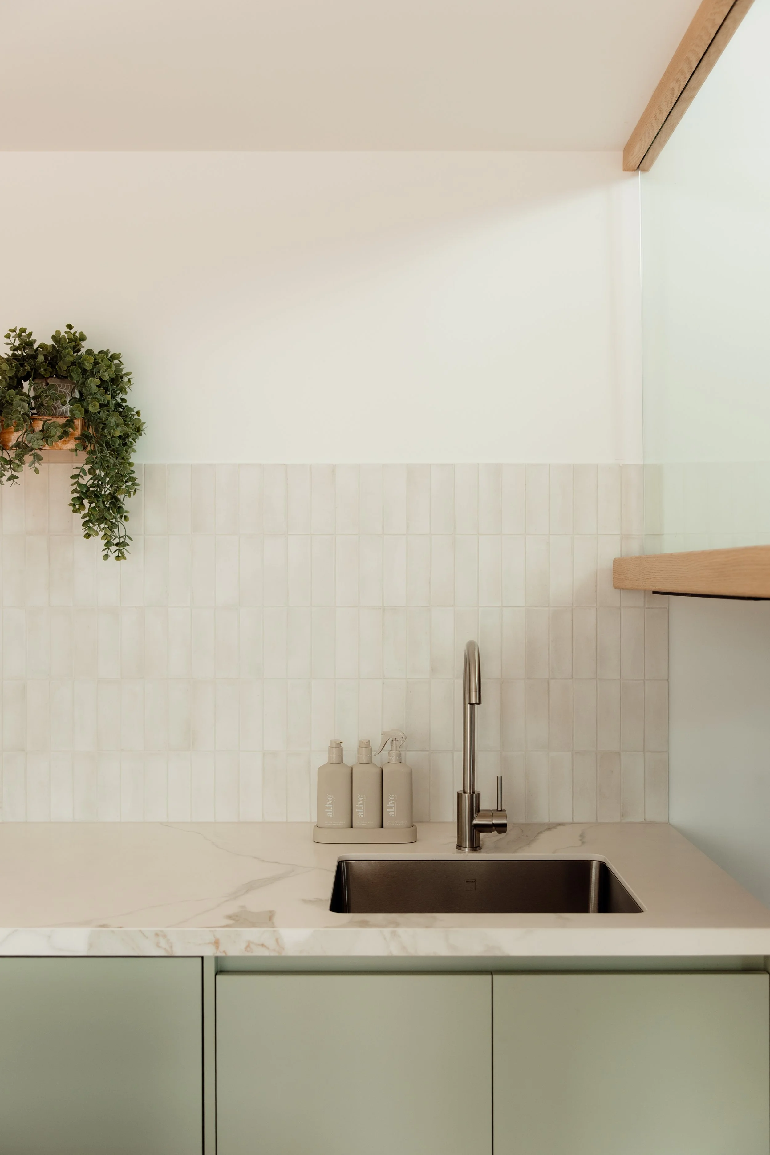 Kitchen sink with beige bottles, a stainless steel faucet, a marble countertop, a light green cabinet, tiled backsplash, and a potted plant on a wooden shelf.