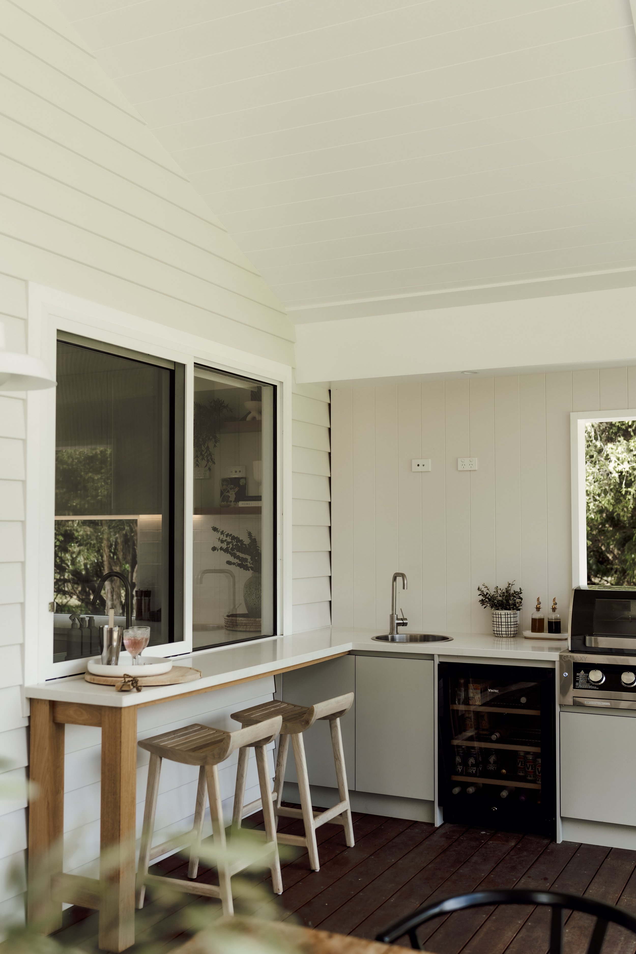 A modern outdoor kitchen with a white countertop, a stainless steel sink, a small wine fridge, and a sliding glass door leading to the indoor space. There are two wooden barstools at a small wooden breakfast bar. The area has white walls and a wooden
