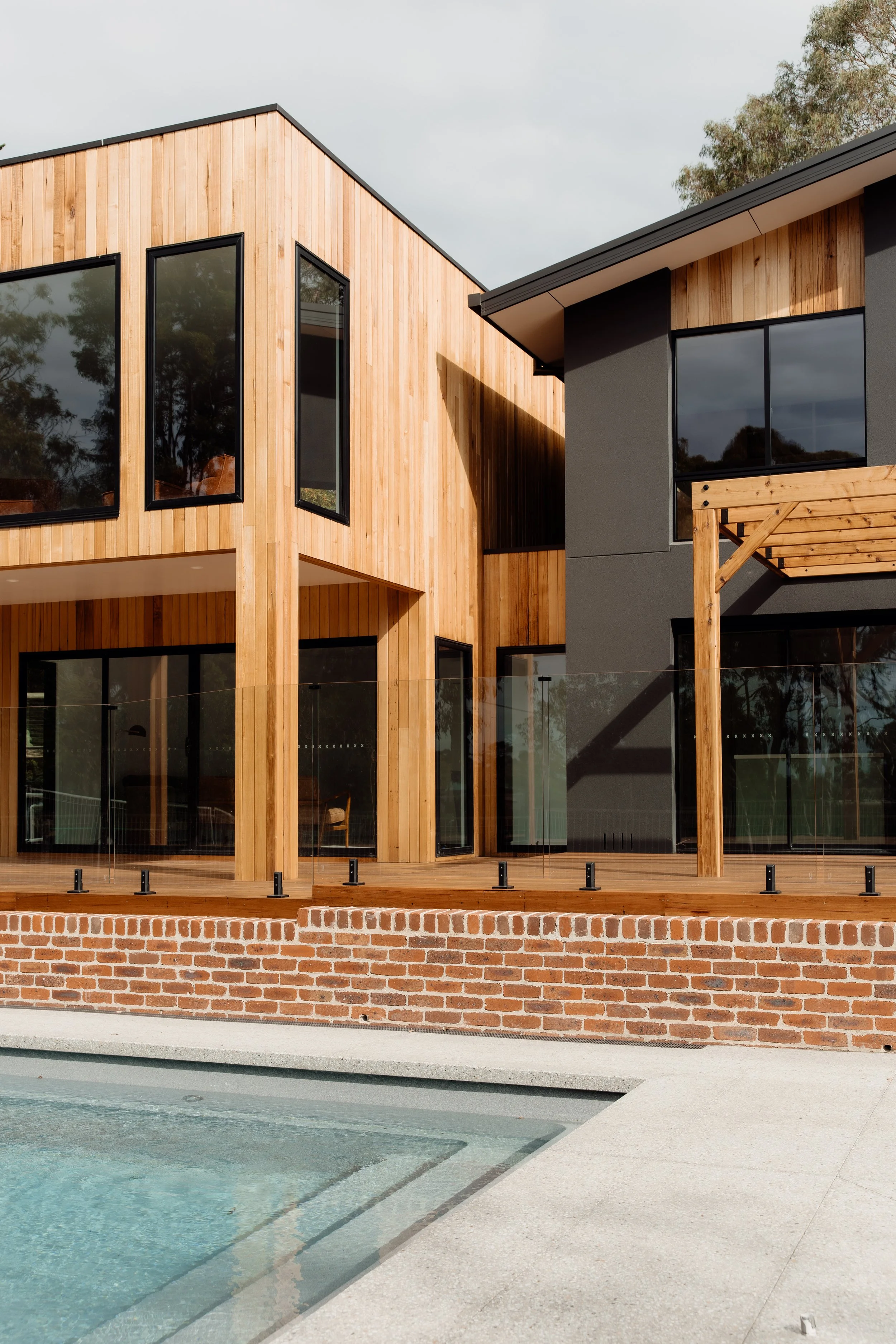 Modern house with wood and gray exterior, large windows, a small balcony, brick foundation, and a swimming pool in the foreground.