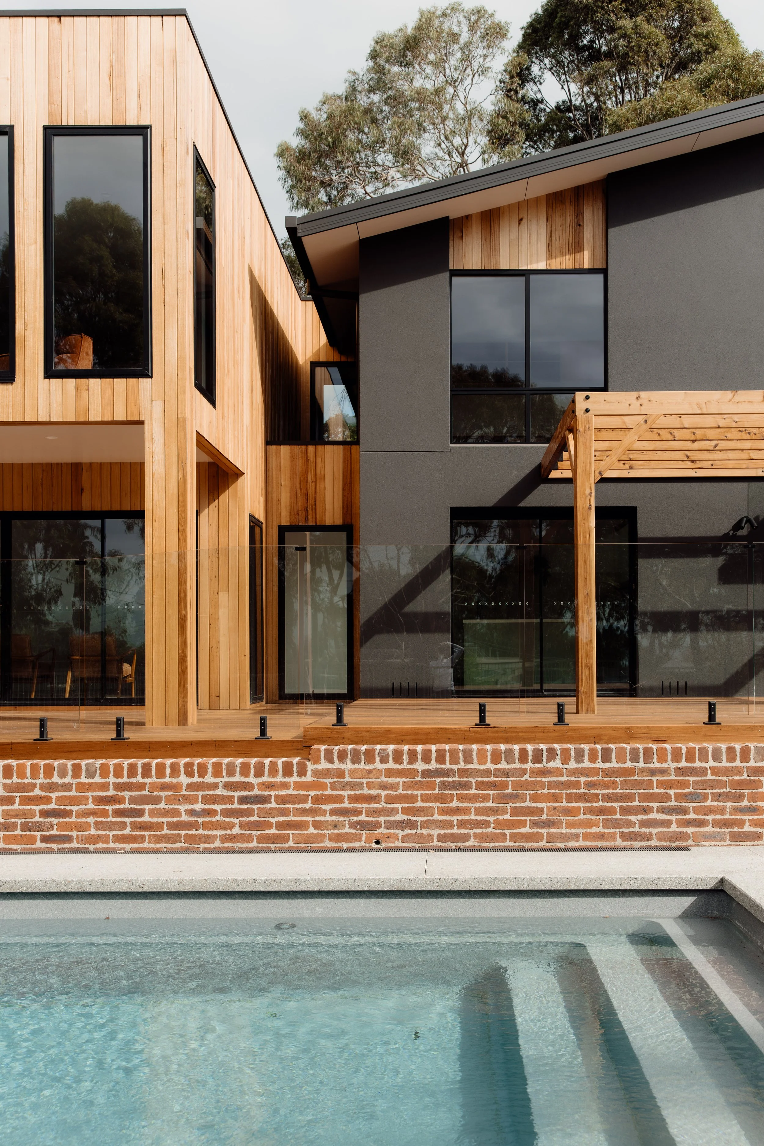 Modern house with a mixed facade of wood and dark gray stucco, large windows, and a wooden deck with glass railing, in front of a swimming pool and trees in the background.