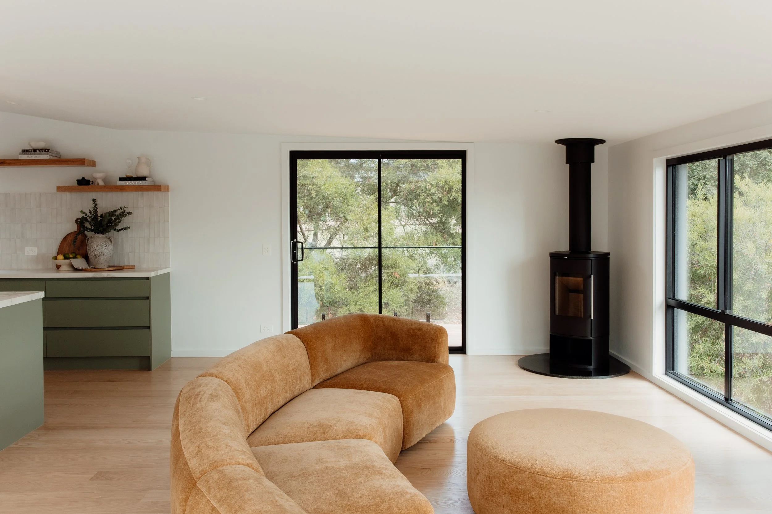 Living room with a curved beige sofa and a matching ottoman facing a large window and a black fireplace stove. There is a sliding glass door leading to an outdoor area, and a corner shelf with decorative items. The room has light wood flooring and mi