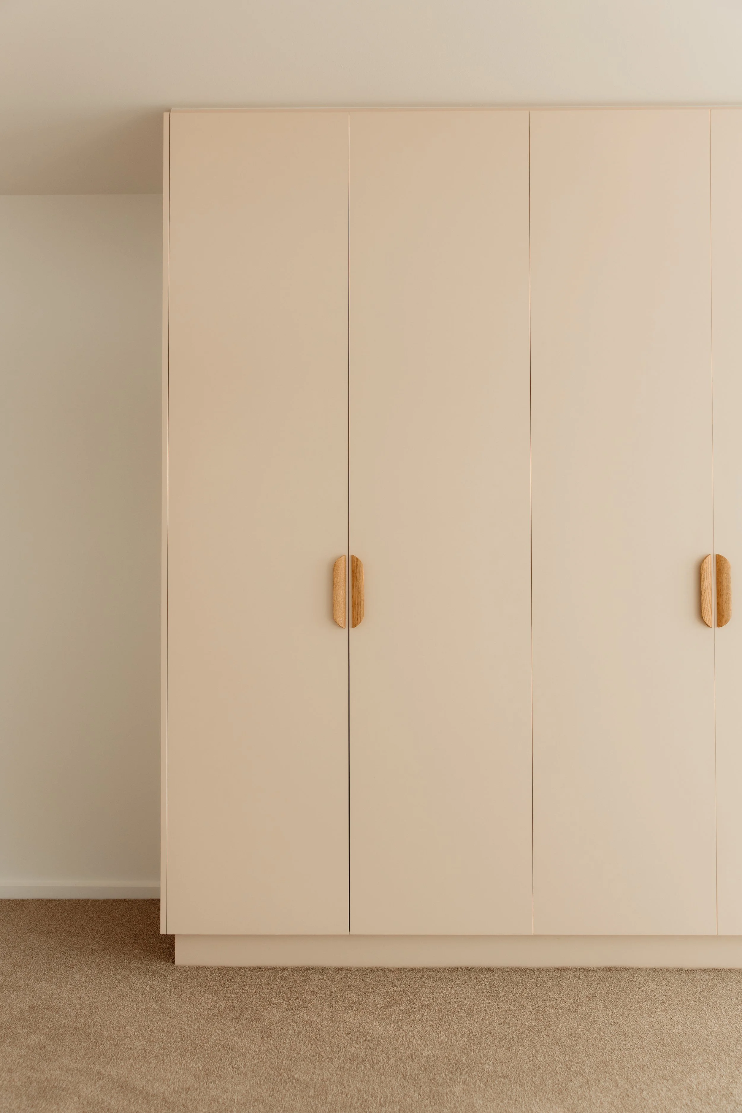 A white wardrobe with four doors and wooden handles, standing on a beige carpet in a room with white walls.