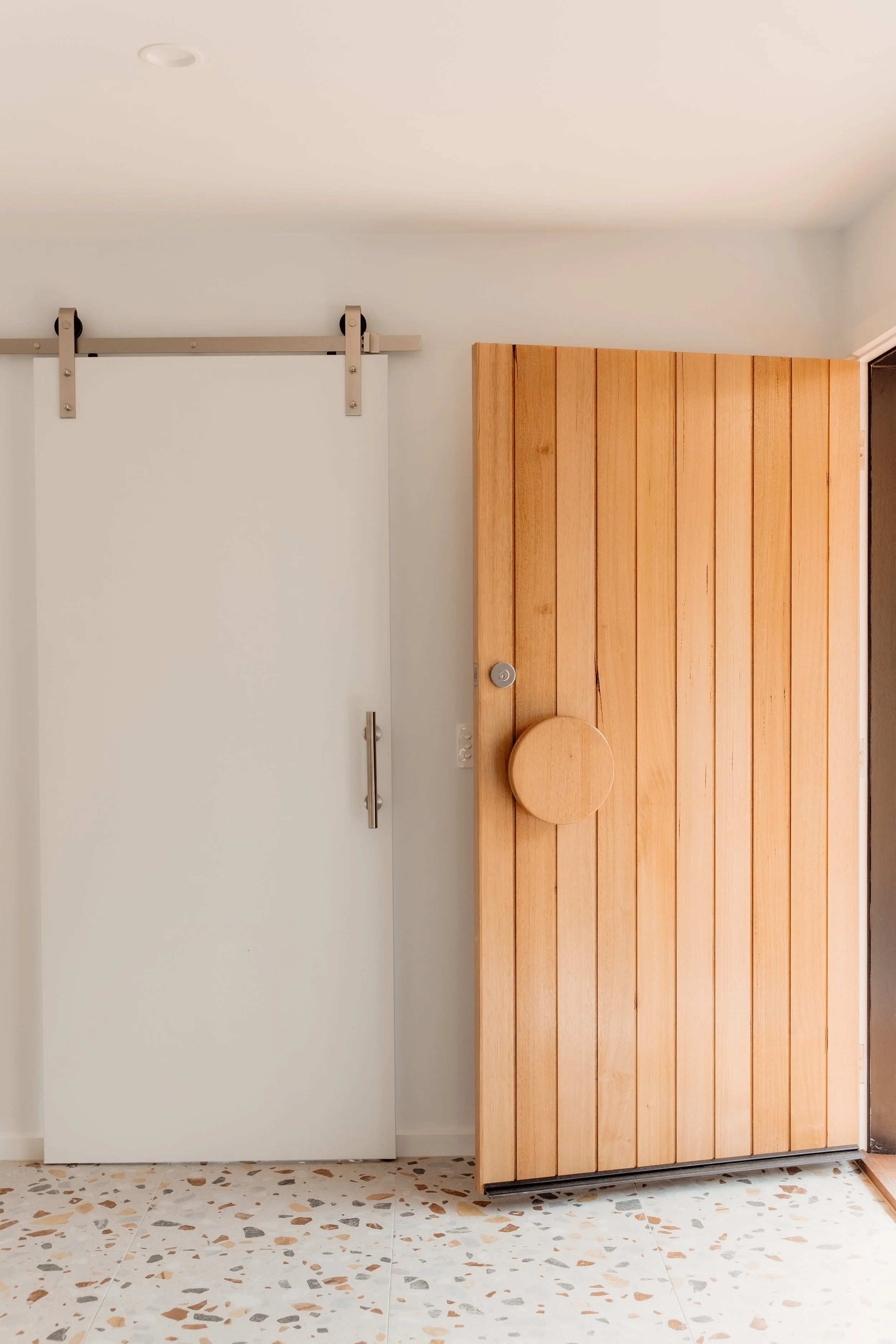 Interior of a modern room featuring a white door with a sliding barn door track above it, a wooden horizontal paneled door, a terrazzo tile floor, and a white wall.