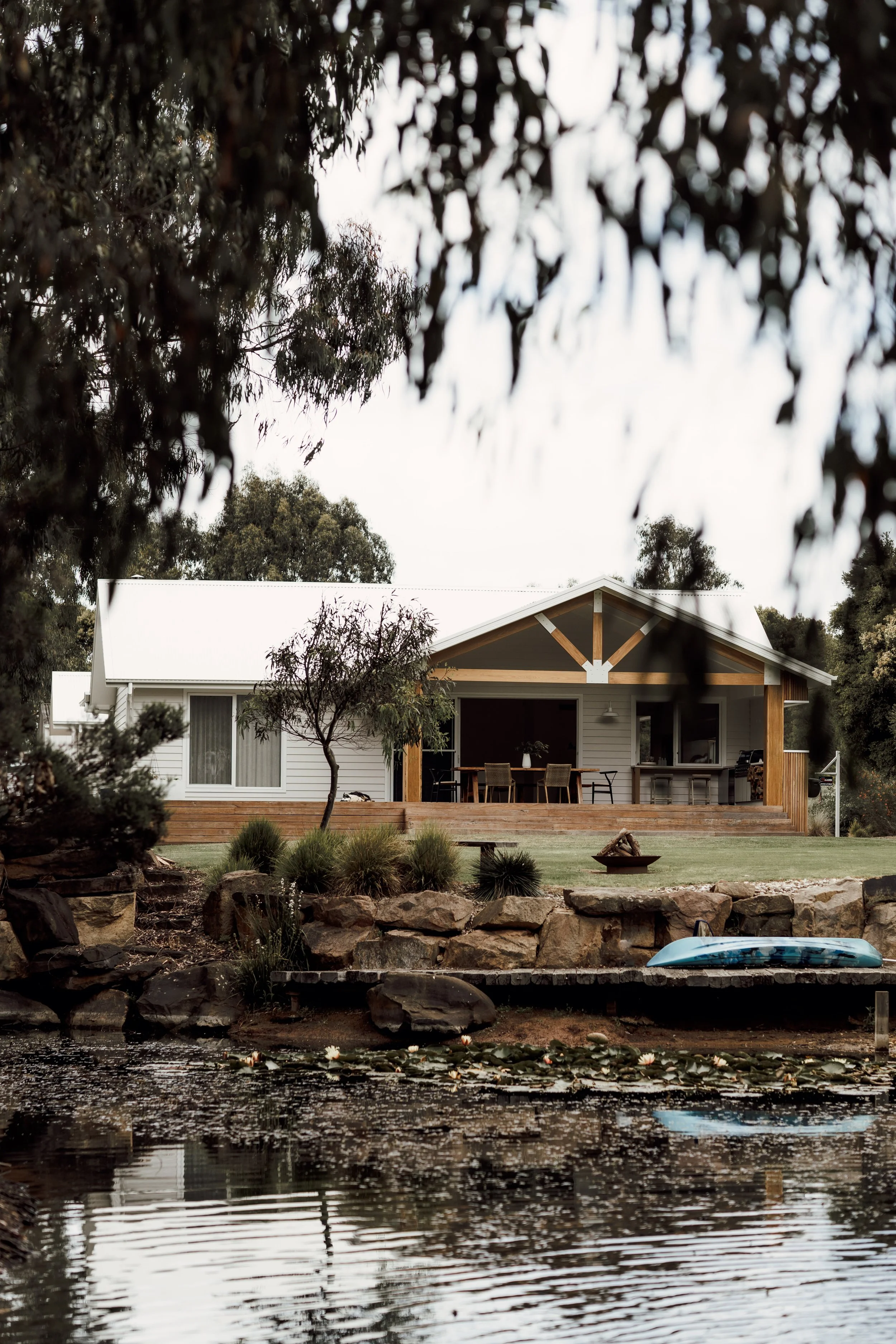 A house with a white exterior and a porch, overlooking a pond with lily pads and a small blue boat docked nearby. Trees surround the house and the sky appears overcast.