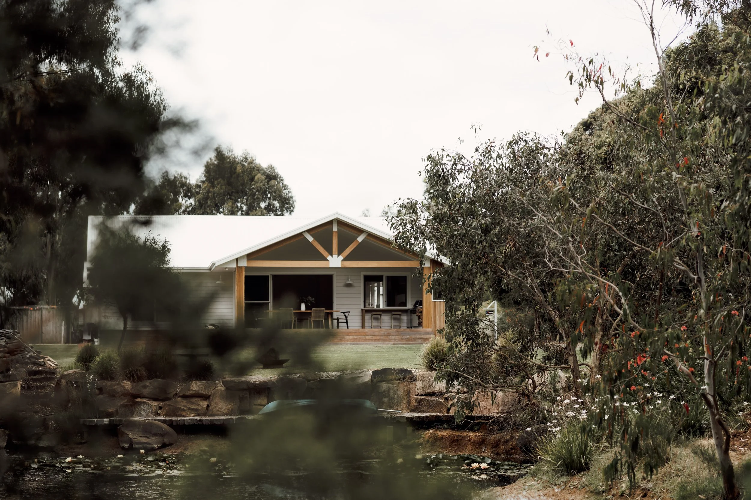 A house with a front porch surrounded by trees and shrubs, with a pond in the foreground.