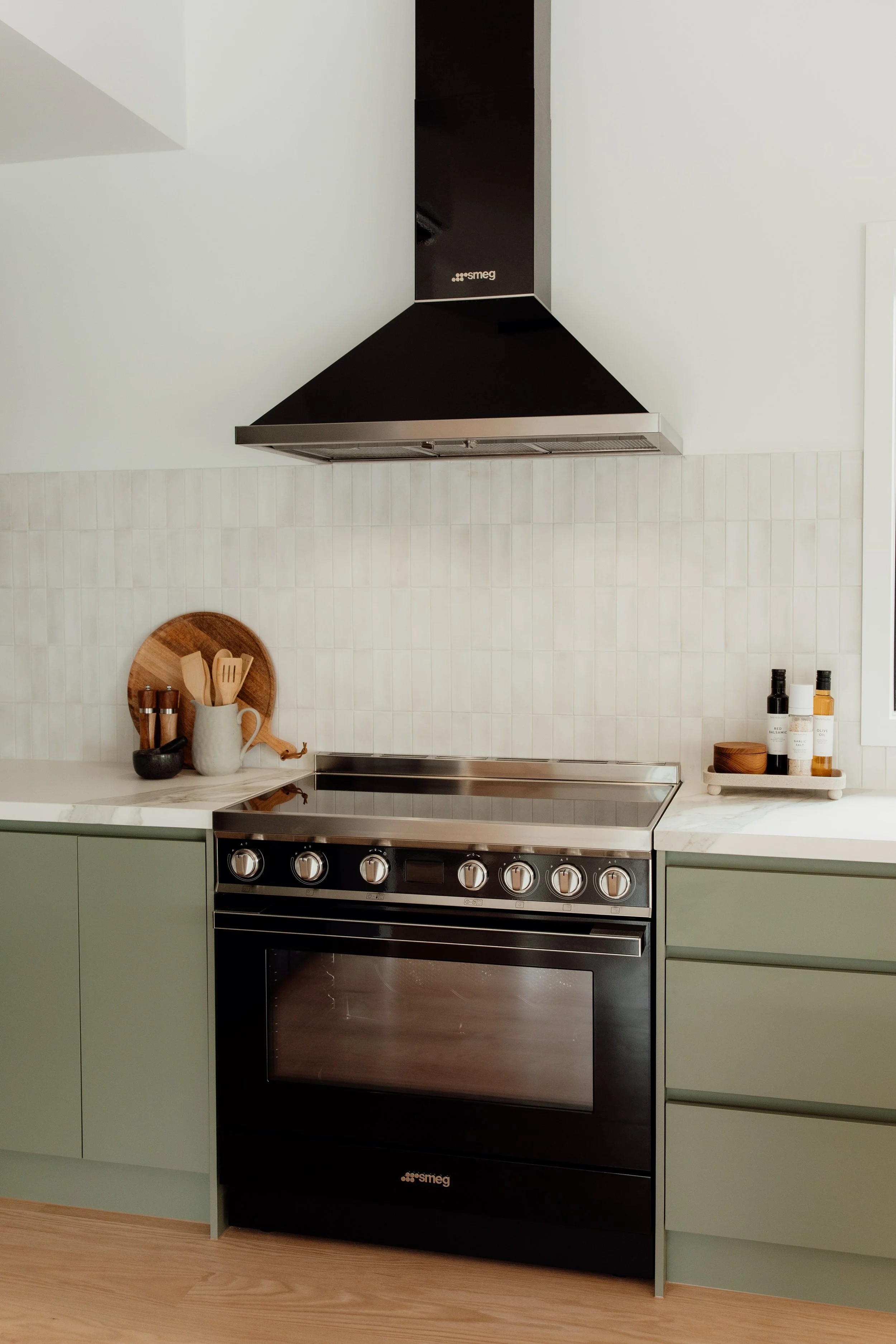 Green kitchen with a black Smeg oven, a marble countertop, and a black range hood. Decorative items include wooden utensils in a white mug, a round wooden tray, and bottles with black caps.