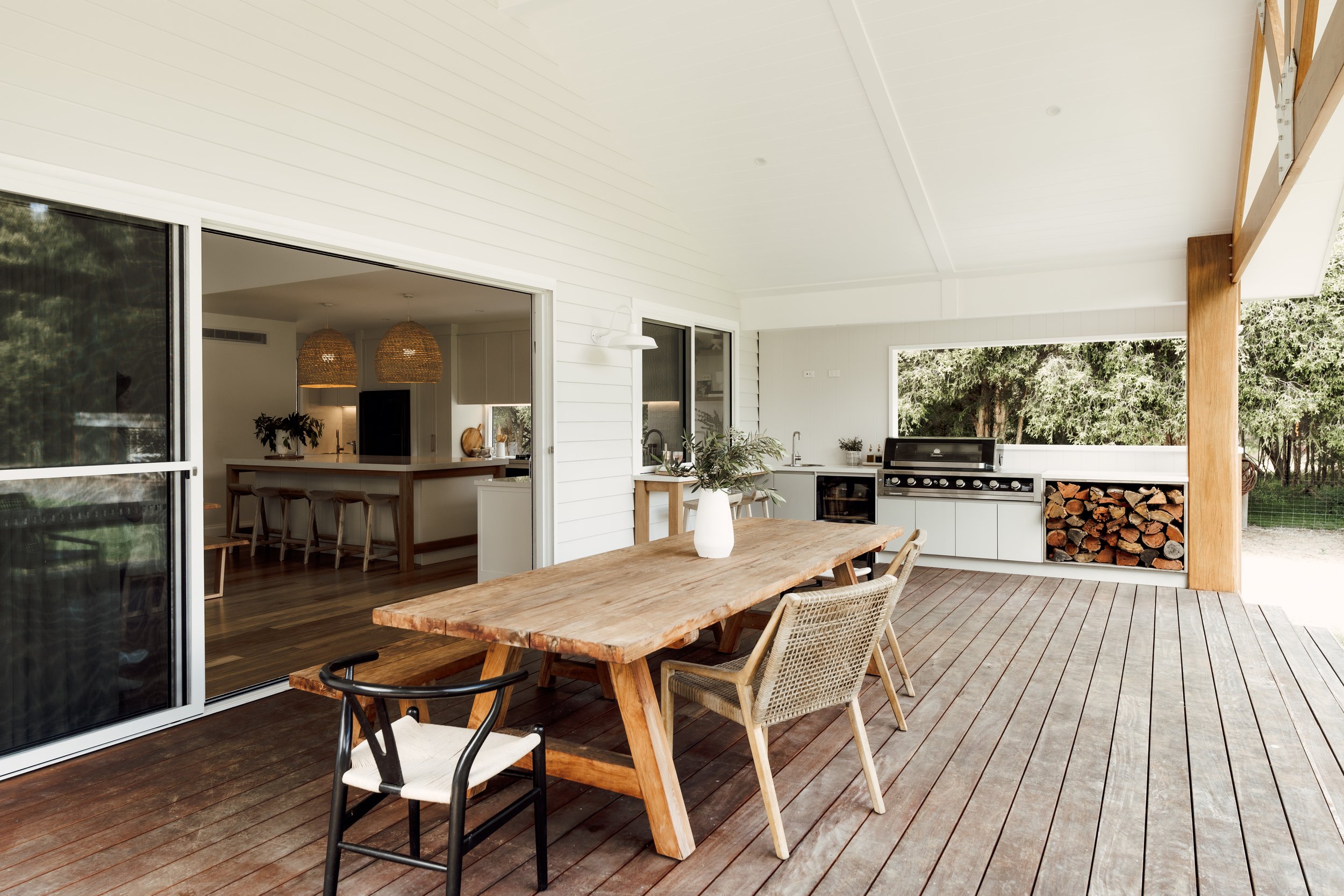 An outdoor patio area with a wooden dining table, chairs, and a kitchen area with a gas stove and firewood storage, adjacent to a house with large sliding glass doors showing an interior kitchen.