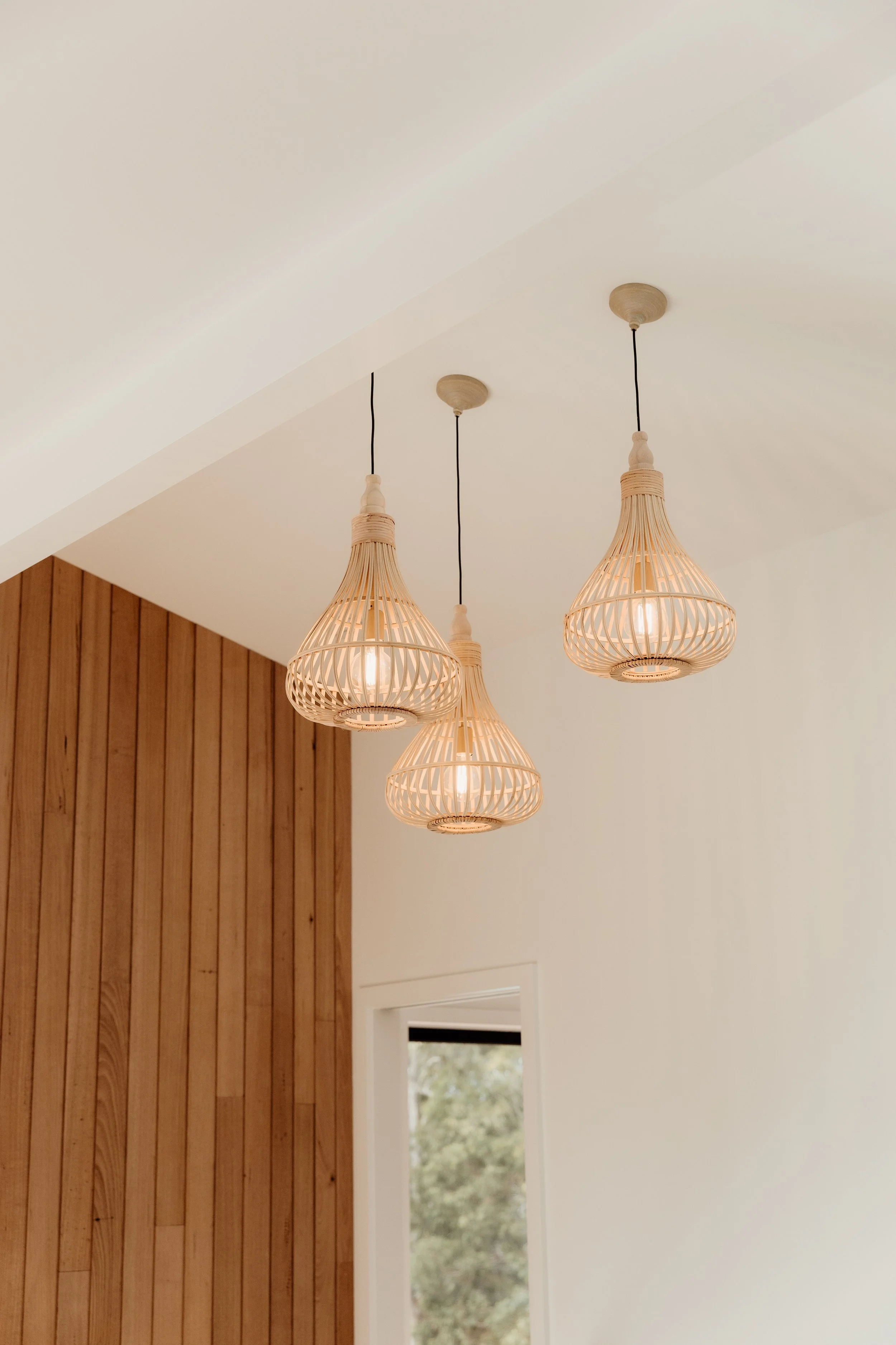 Three hanging pendant lights with wooden slatted shades in a room with a wooden wall and a window showing greenery outside.
