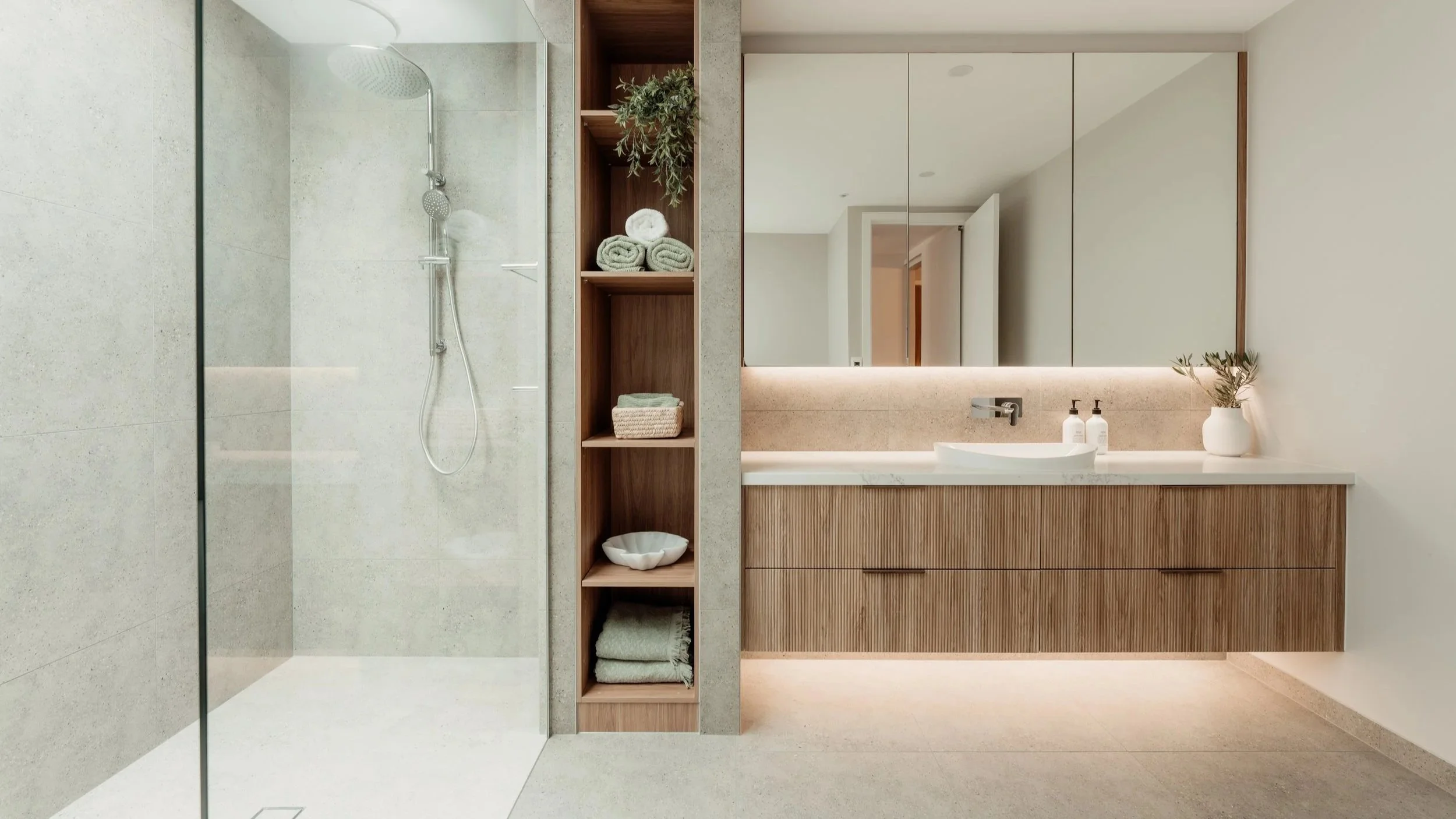 Modern bathroom with a walk-in shower on the left, open shelving with rolled towels and decorative items in the middle, large mirror with cabinet doors above the sink on the right, beige and wood tones, potted plant on the sink
