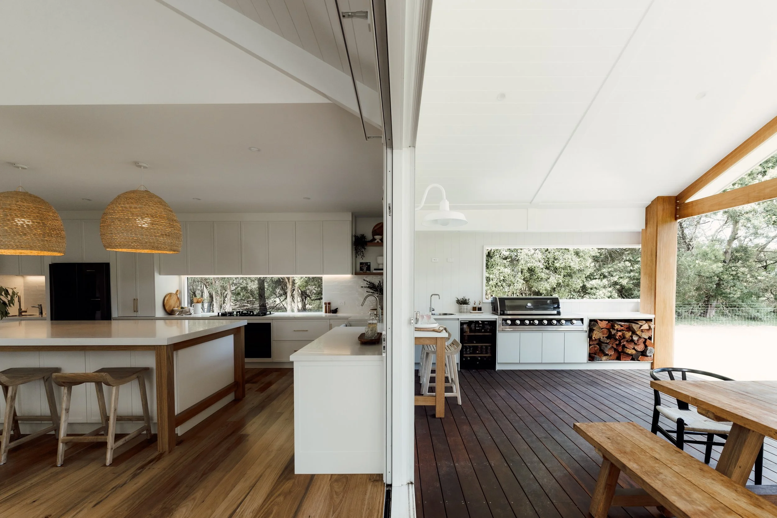The image shows a split view of a modern kitchen and outdoor patio. The kitchen has a white island, hanging woven pendant lights, and white cabinets. The outdoor patio features a wooden table, chairs, and a wood-burning stove with stacked firewood un