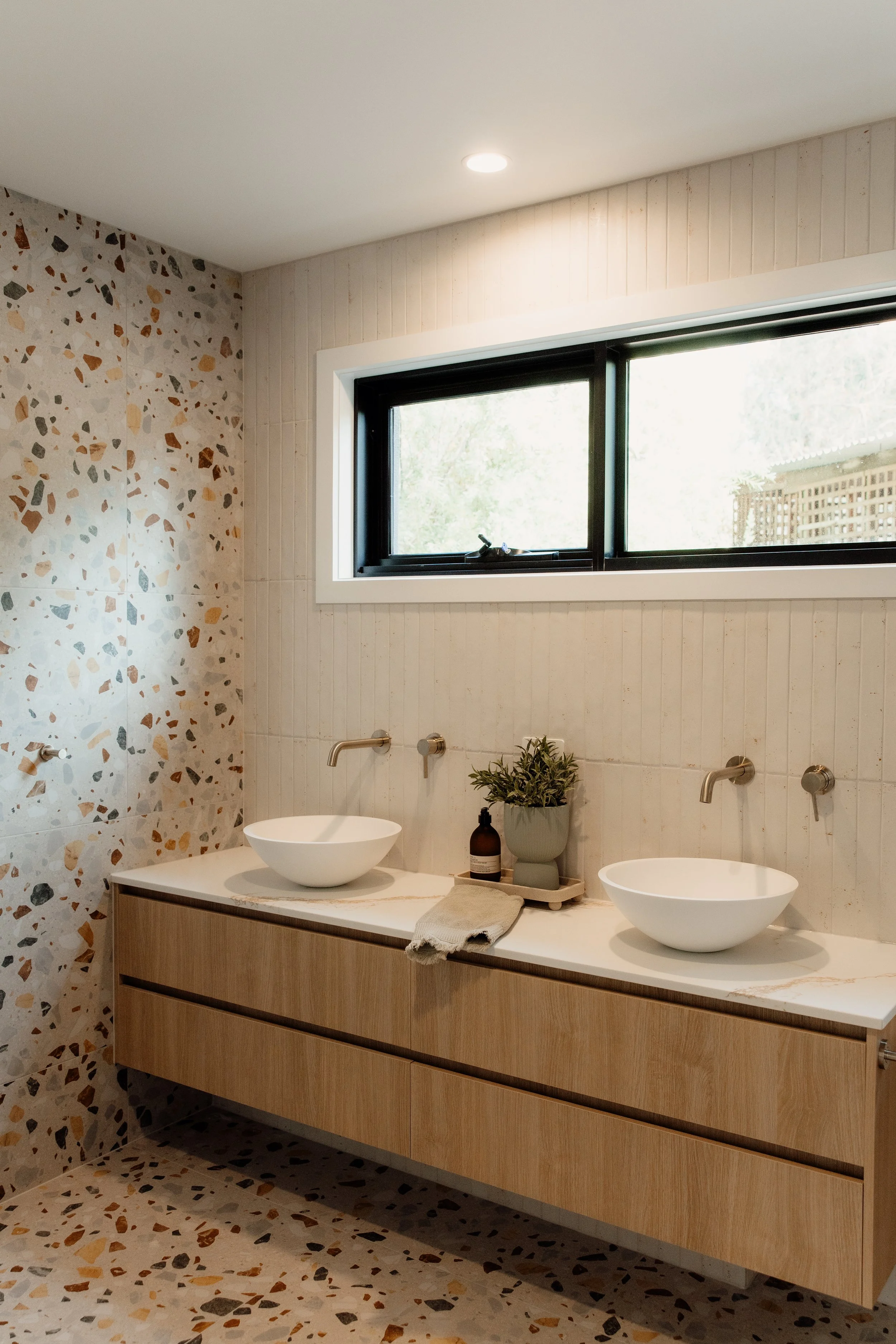 Modern bathroom with terrazzo tiles, a large window, and a wooden vanity with two vessel sinks, a plant, and toiletries.