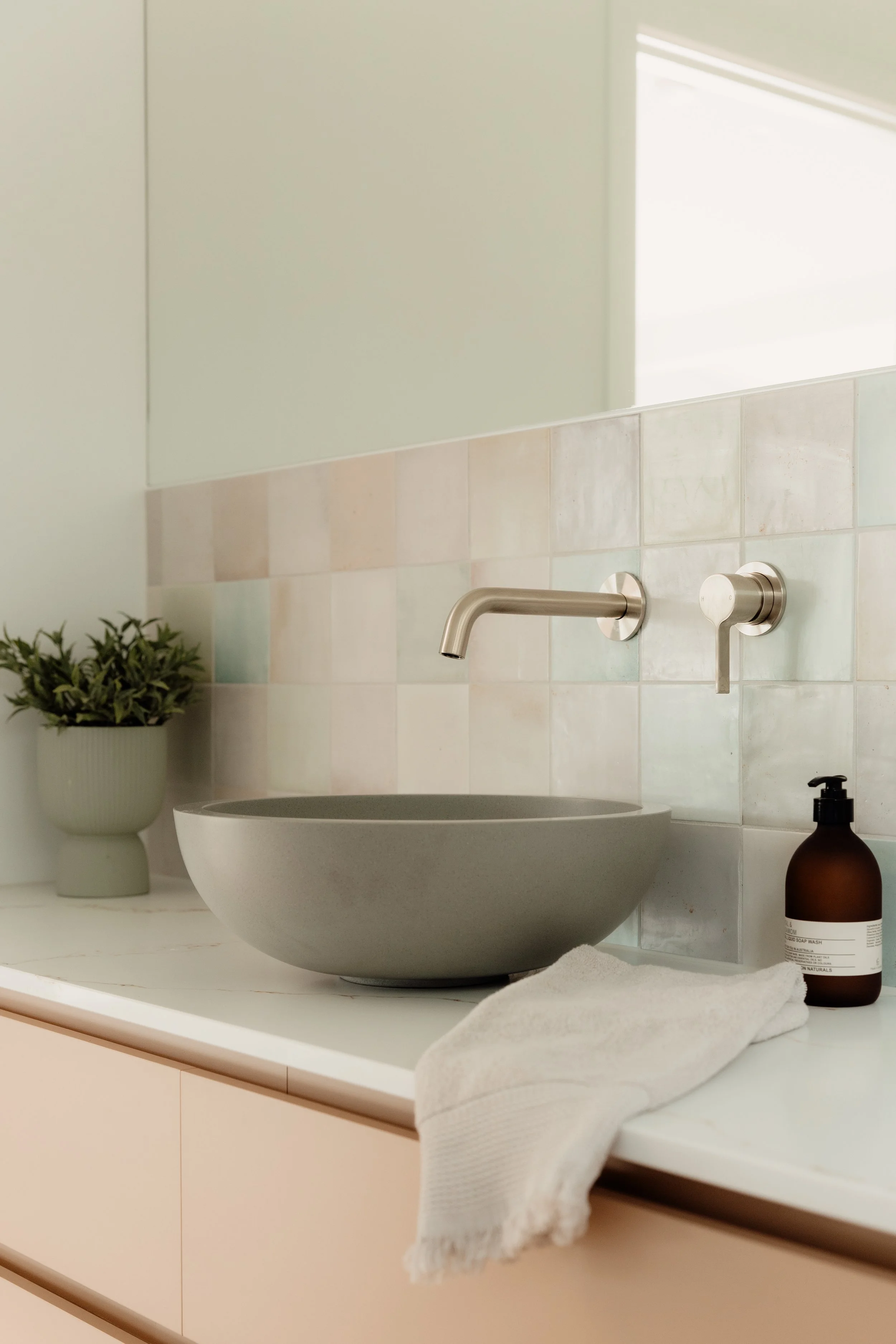 Bathroom sink with a beige bowl-shaped vessel, a matte black soap dispenser, and a white towel on a white countertop, with a potted plant nearby and a wall mirror above.