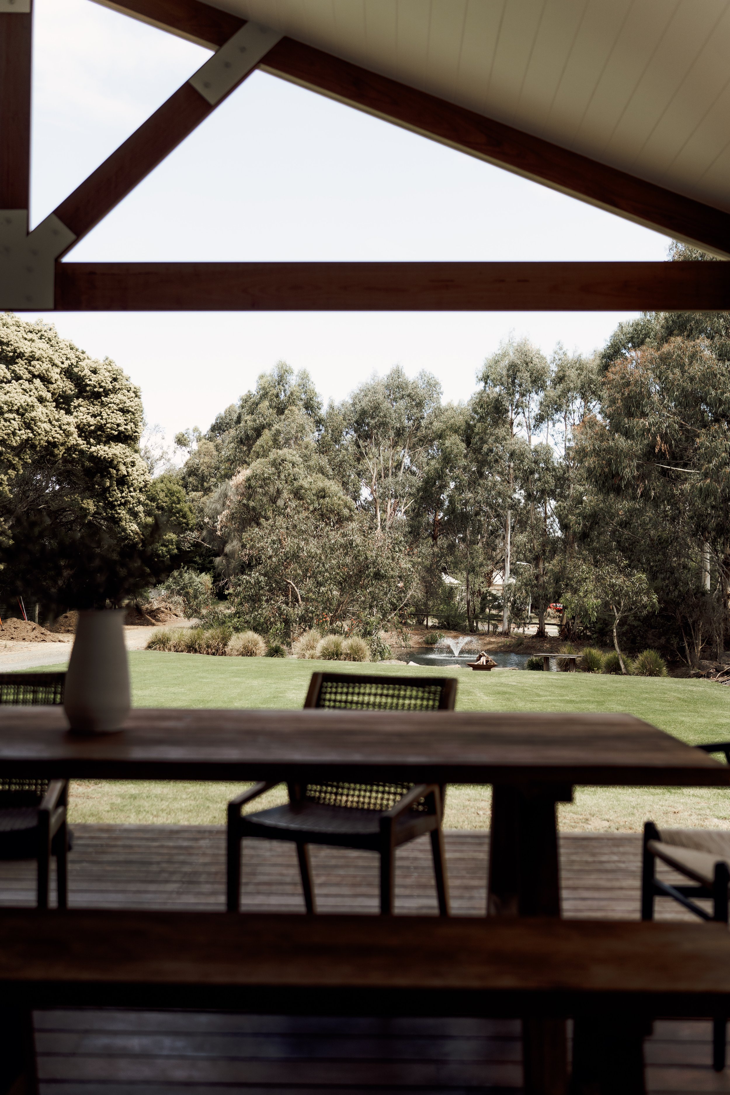 View from under a covered patio or porch overlooking a backyard with green grass, trees, and a pond with a fountain.