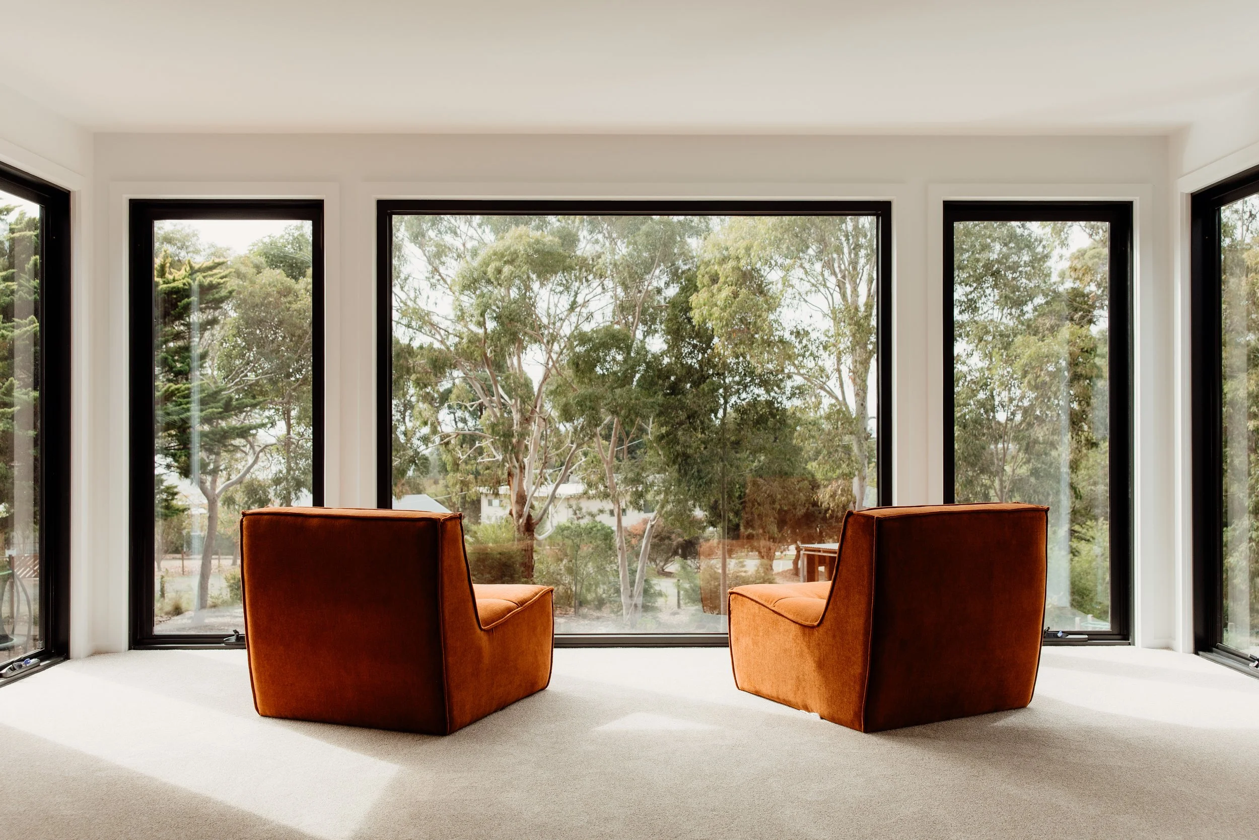Modern room with large floor-to-ceiling windows overlooking trees and a suburban neighborhood; two orange upholstered chairs facing the windows.