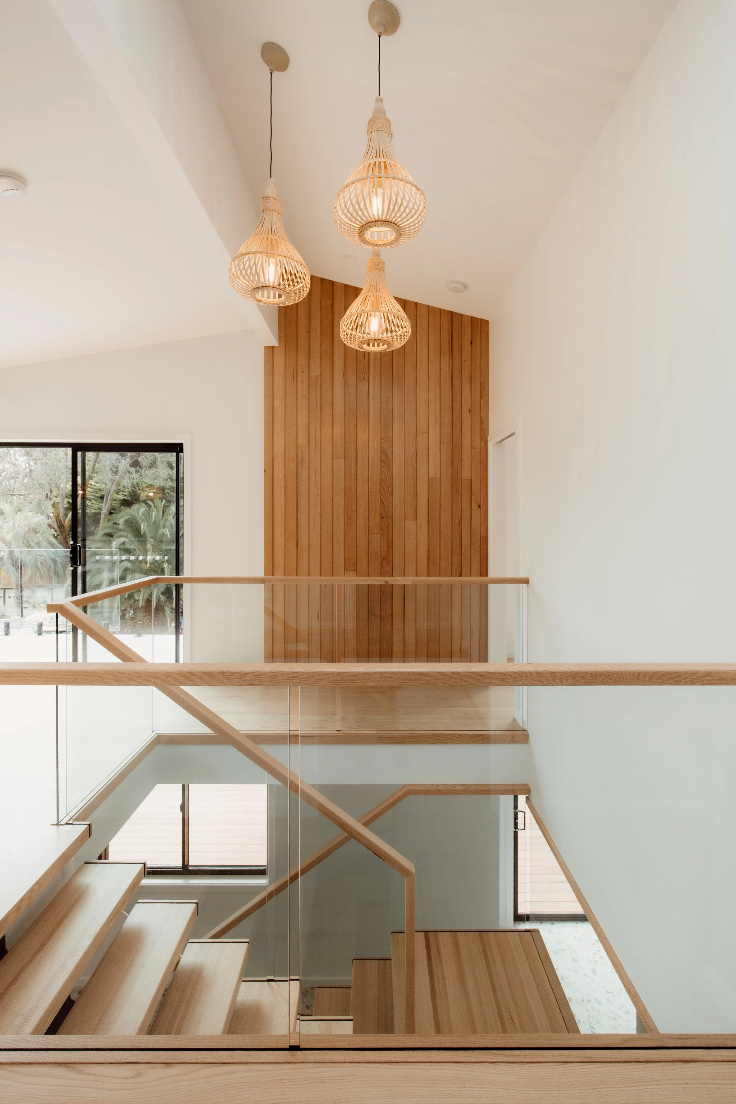 Interior of a modern home featuring a staircase with wood and glass railing, white walls, wooden wall panel, and three pendant lights hanging from the ceiling.