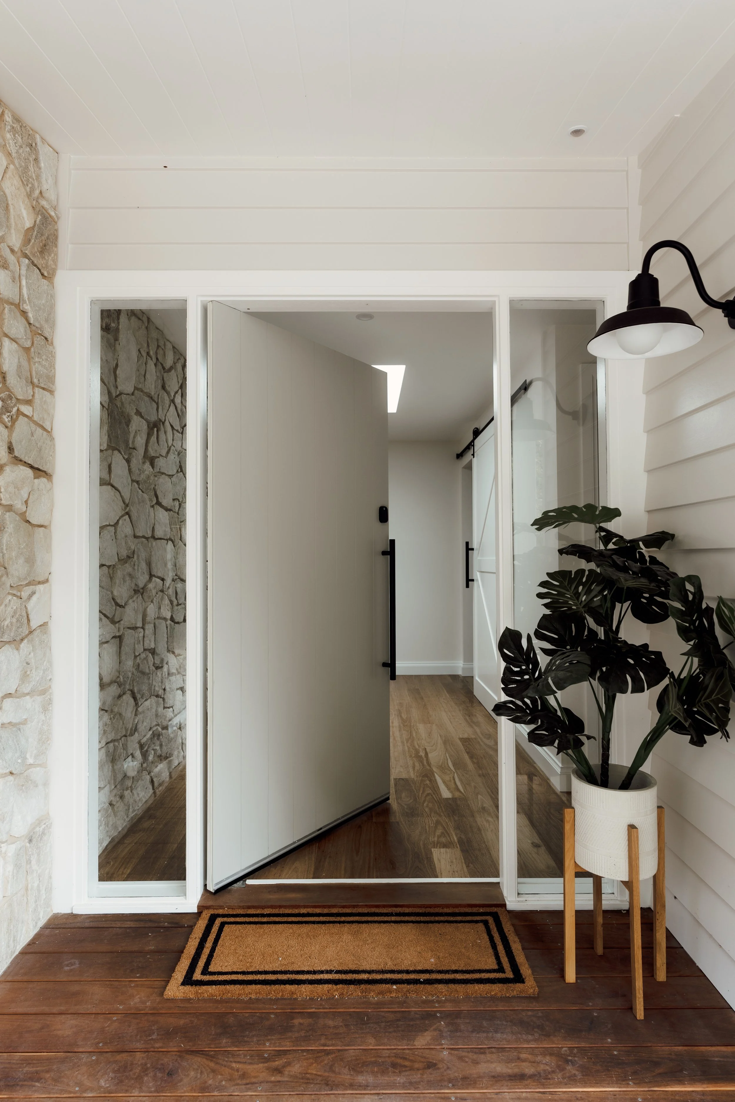 Modern interior entrance with wooden floor, indoor plants on a stand, and a sliding glass door leading to a hallway with a barn door and hardwood floor.