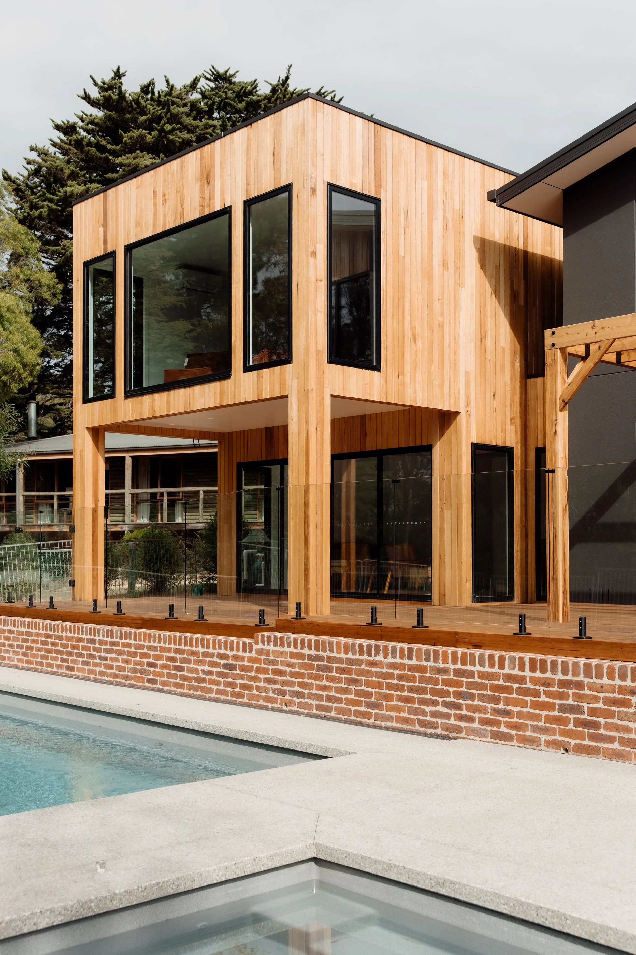 Modern building with wooden exterior and large black-framed windows, located near a swimming pool with a brick boundary.