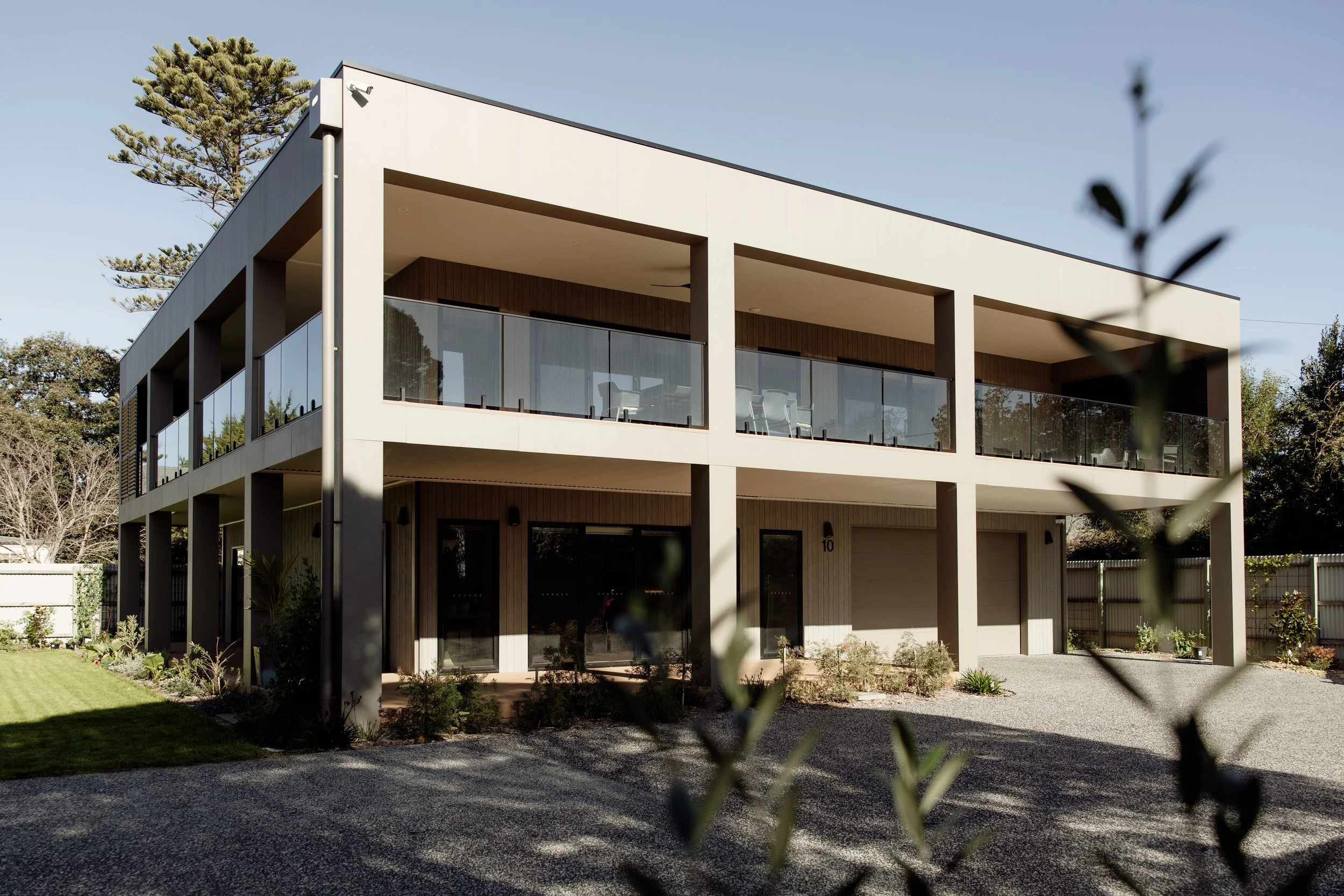 Modern two-story house with large glass balconies, surrounded by trees and a gravel driveway, under a clear blue sky.